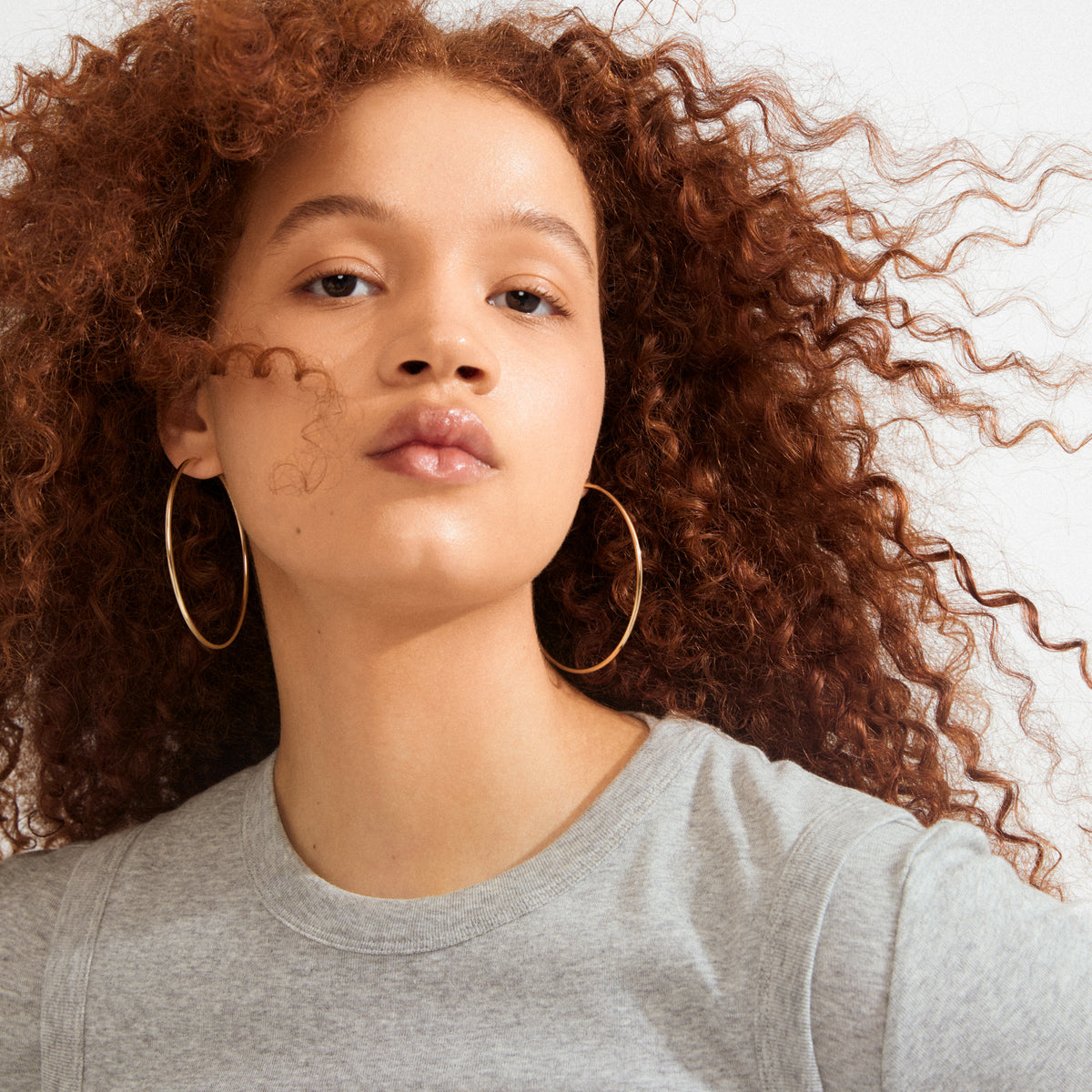 Young woman wearing APRIL large gold-plated hoop earrings, showcasing their sleek, stylish design. Her reddish-brown curls frame the elegant, recycled hoops beautifully.