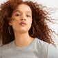 Young woman wearing APRIL large gold-plated hoop earrings, showcasing their sleek, stylish design. Her reddish-brown curls frame the elegant, recycled hoops beautifully.