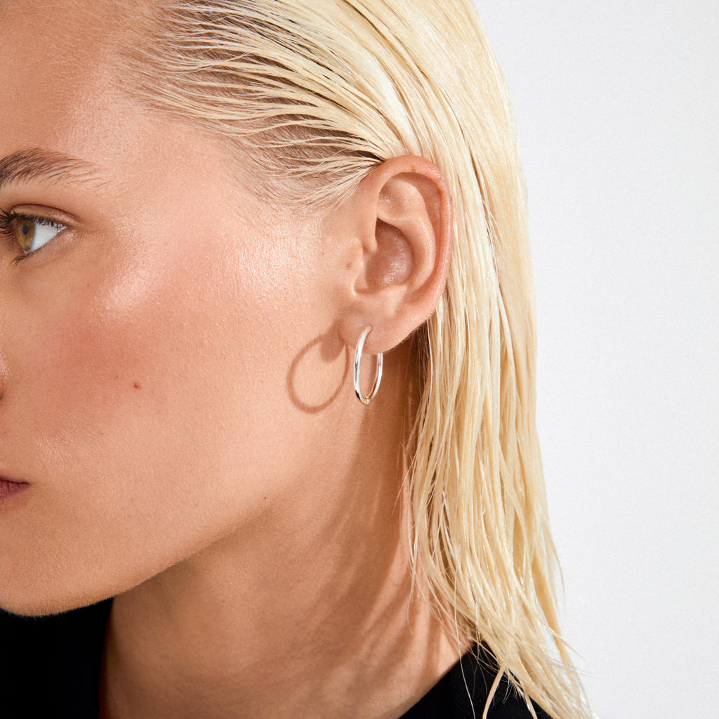 Close-up of APRIL small hoop earrings in silver-plated design, worn on a model's ear with sleek blonde hair, highlighting the earring's polished, timeless appeal.