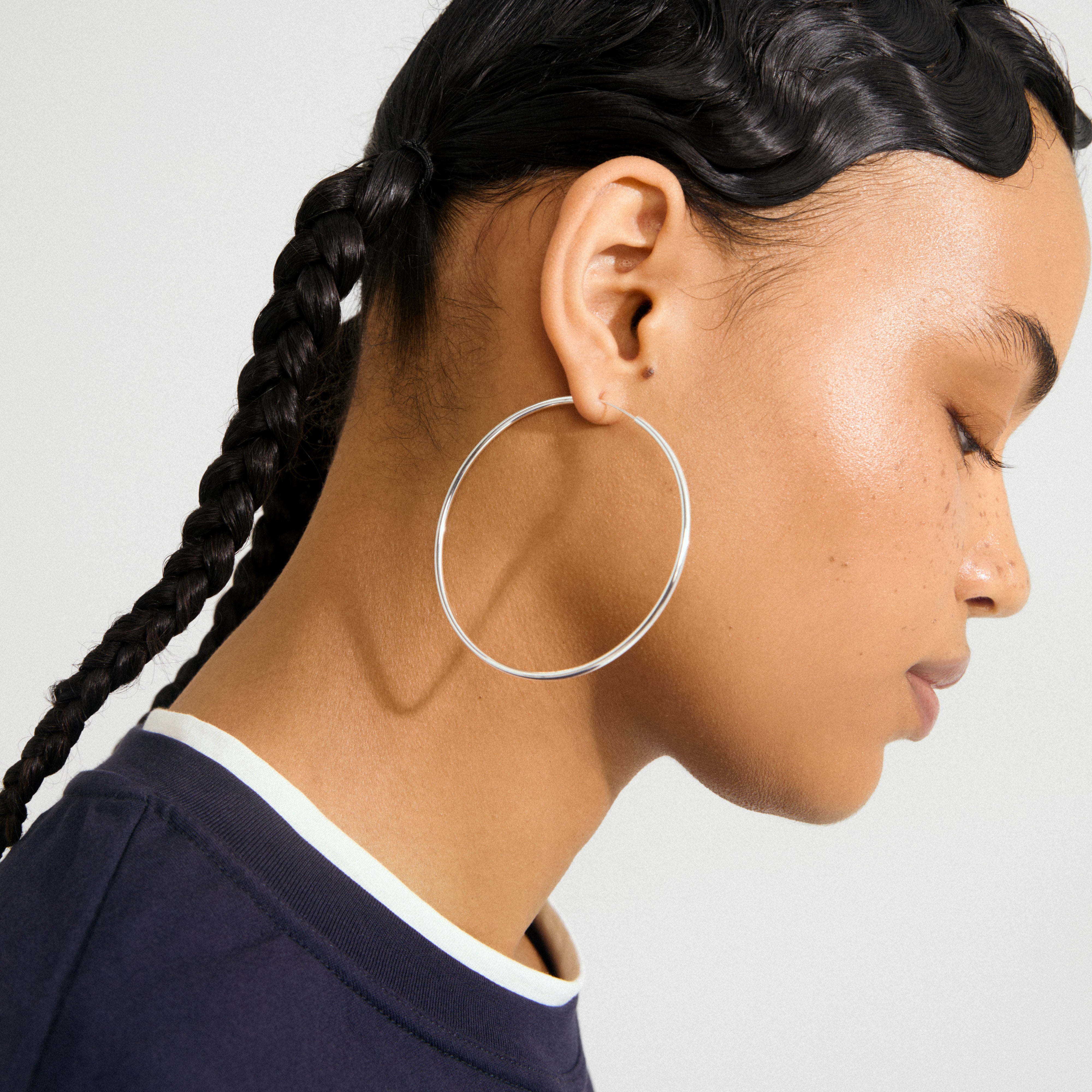 Side profile showcasing APRIL large silver-plated hoop earring, 60 mm diameter, highlighting elegant design with recycled material, worn by a young woman with braided hair.