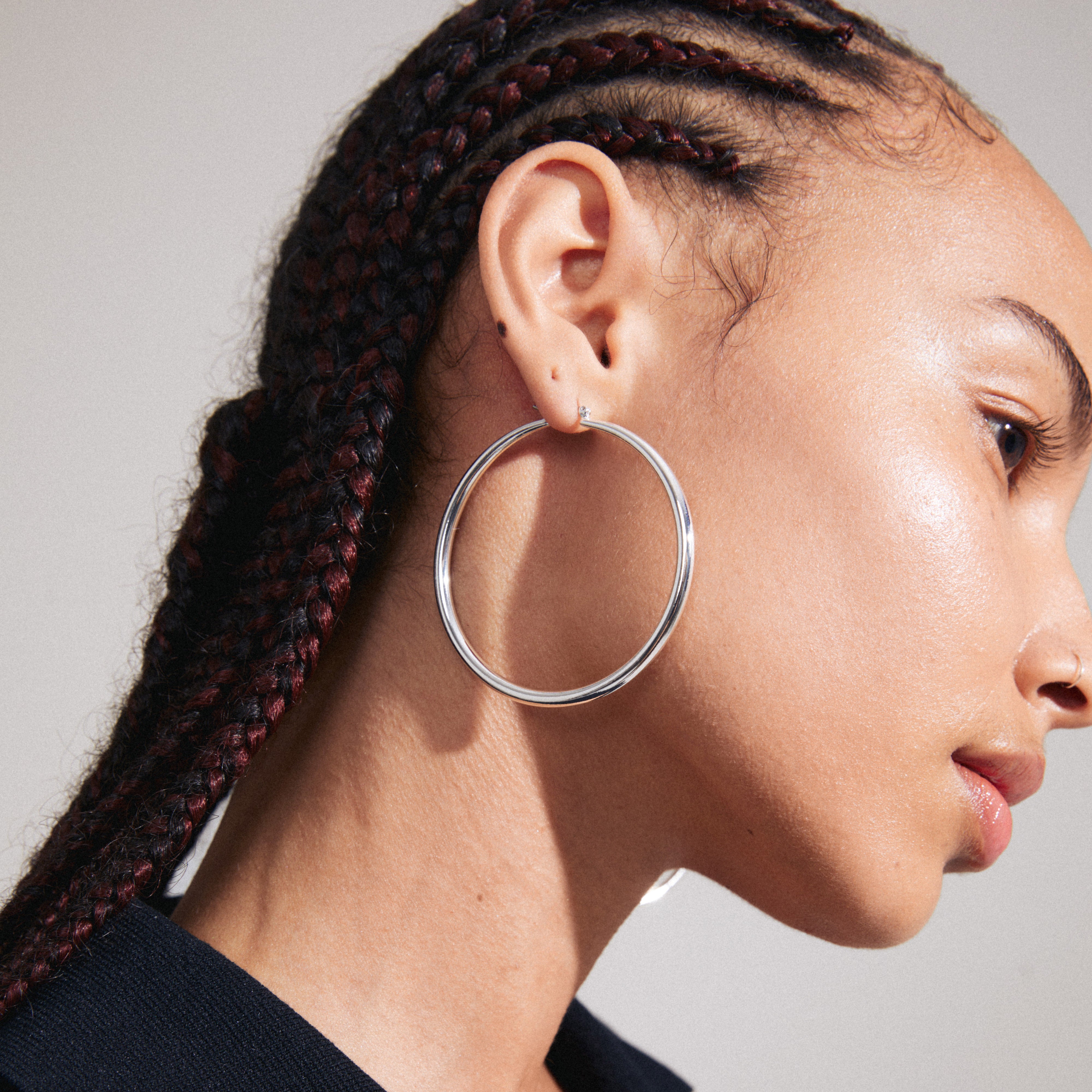 PRIYA large silver-plated hoop earrings showcased on a woman with braided hair, highlighting their classic 60mm size and shiny finish.