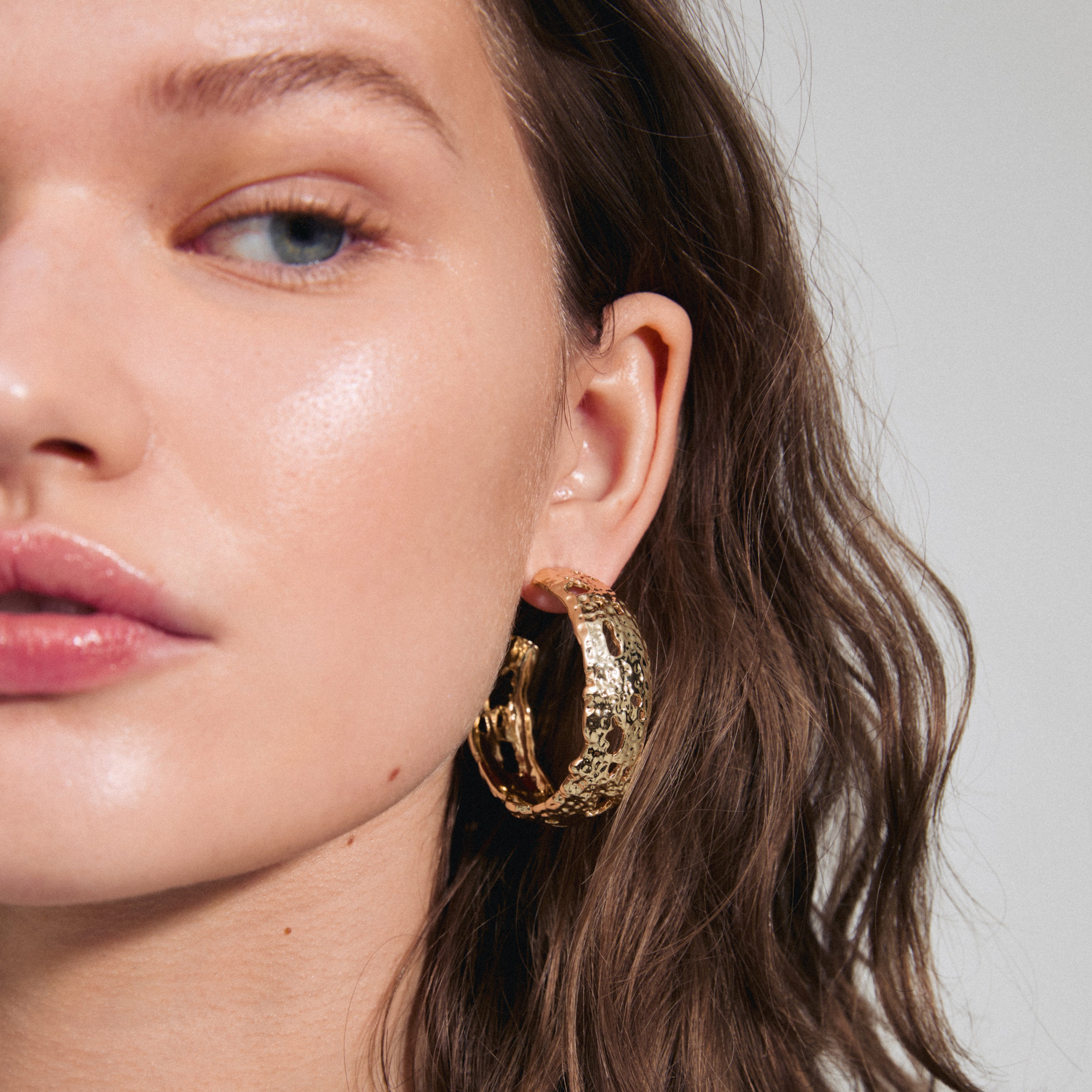 Close-up of a woman wearing Pilgrim's ELENI earrings, showcasing large, gold-plated hoops with a rustic design, emphasizing unique style and eco-friendly craftsmanship.