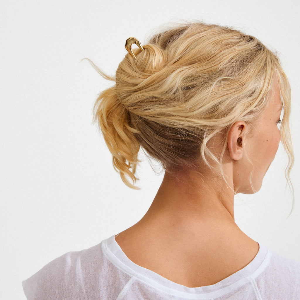 Blonde hair styled in a low bun with the gold-plated AMARIS hairpin from Pilgrim, showcasing its sleek design and chic elegance for versatile hairstyles.