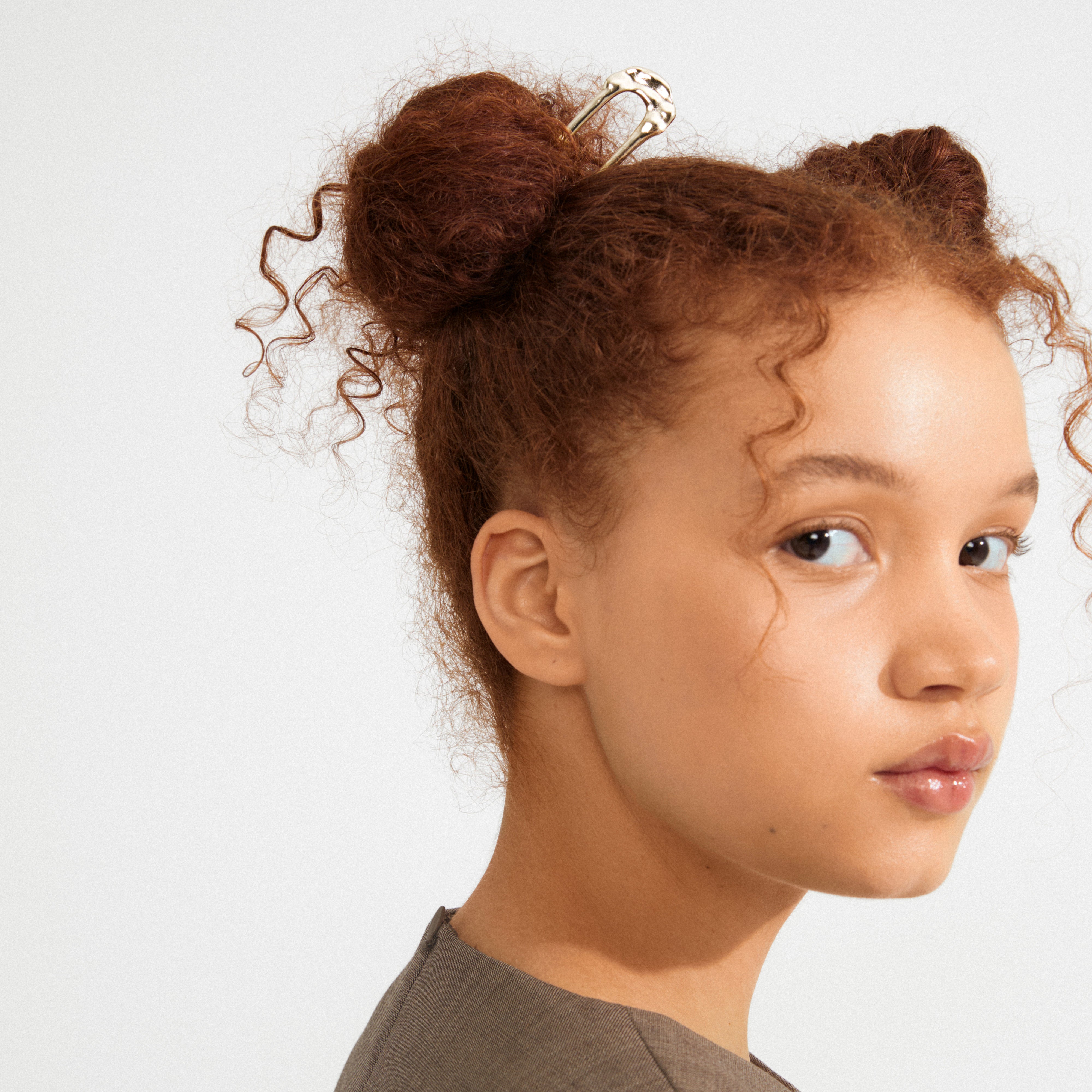 Young person with curly hair styled in buns, adorned with Pilgrim’s gold-plated AMARIS hairpin, showcasing its sleek, minimalist design and understated luxury.