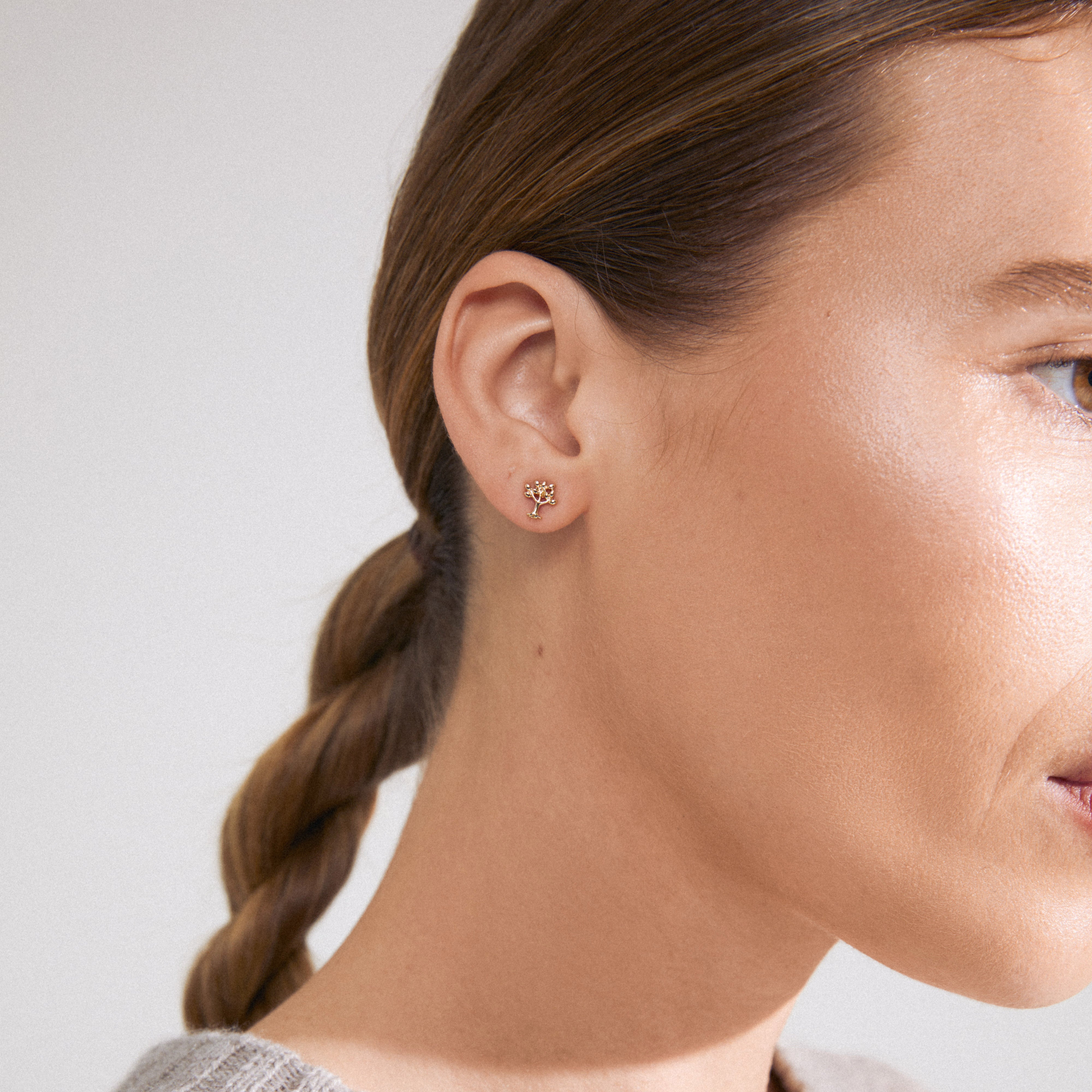 Close-up of a woman's ear wearing the gold-plated PHYLLIS single earring, shaped like the tree of life, symbolizing growth and connection.
