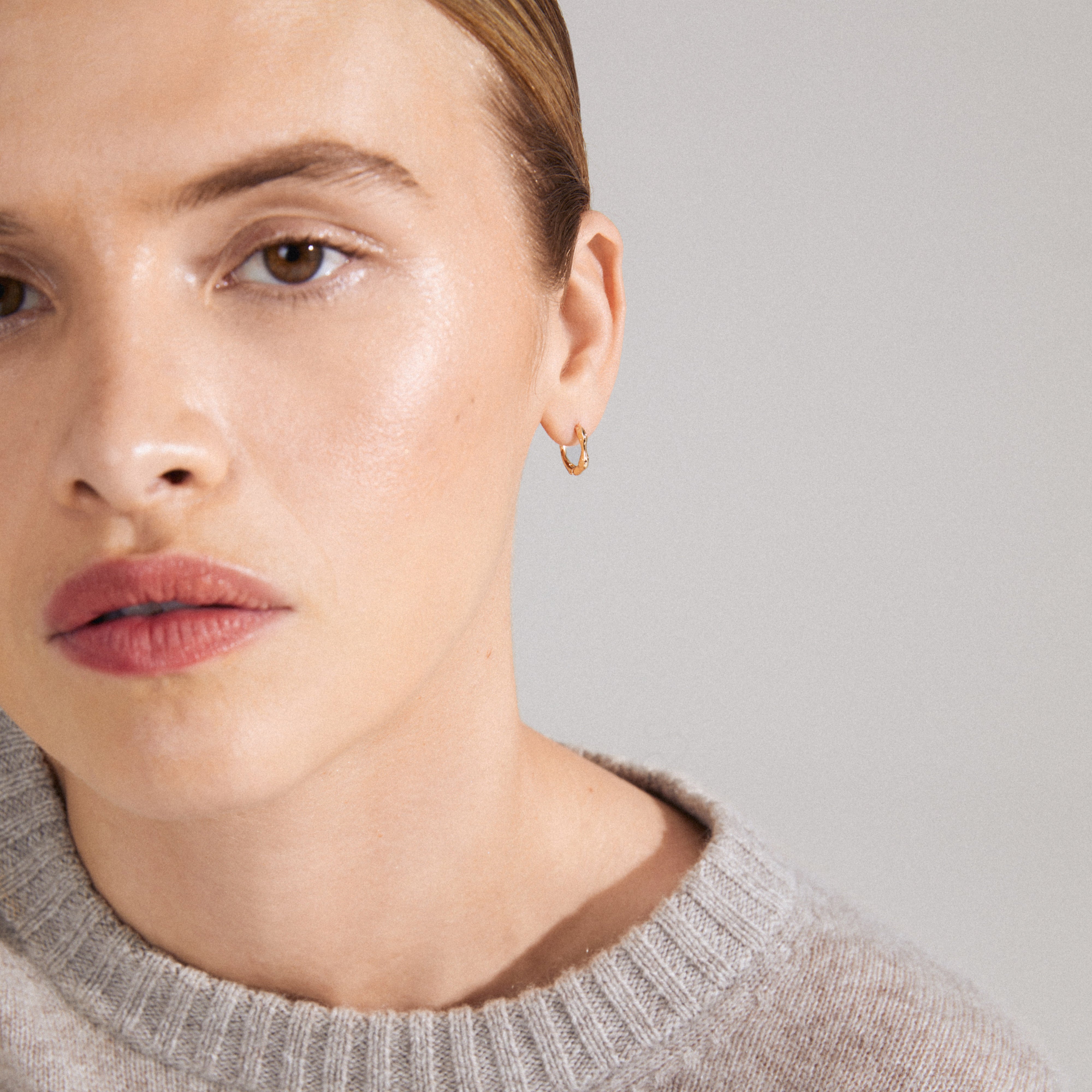 KYRA single earring gold-plated with an organic shape and shiny finish, worn by a woman in a grey sweater, showcasing its handcrafted elegance.