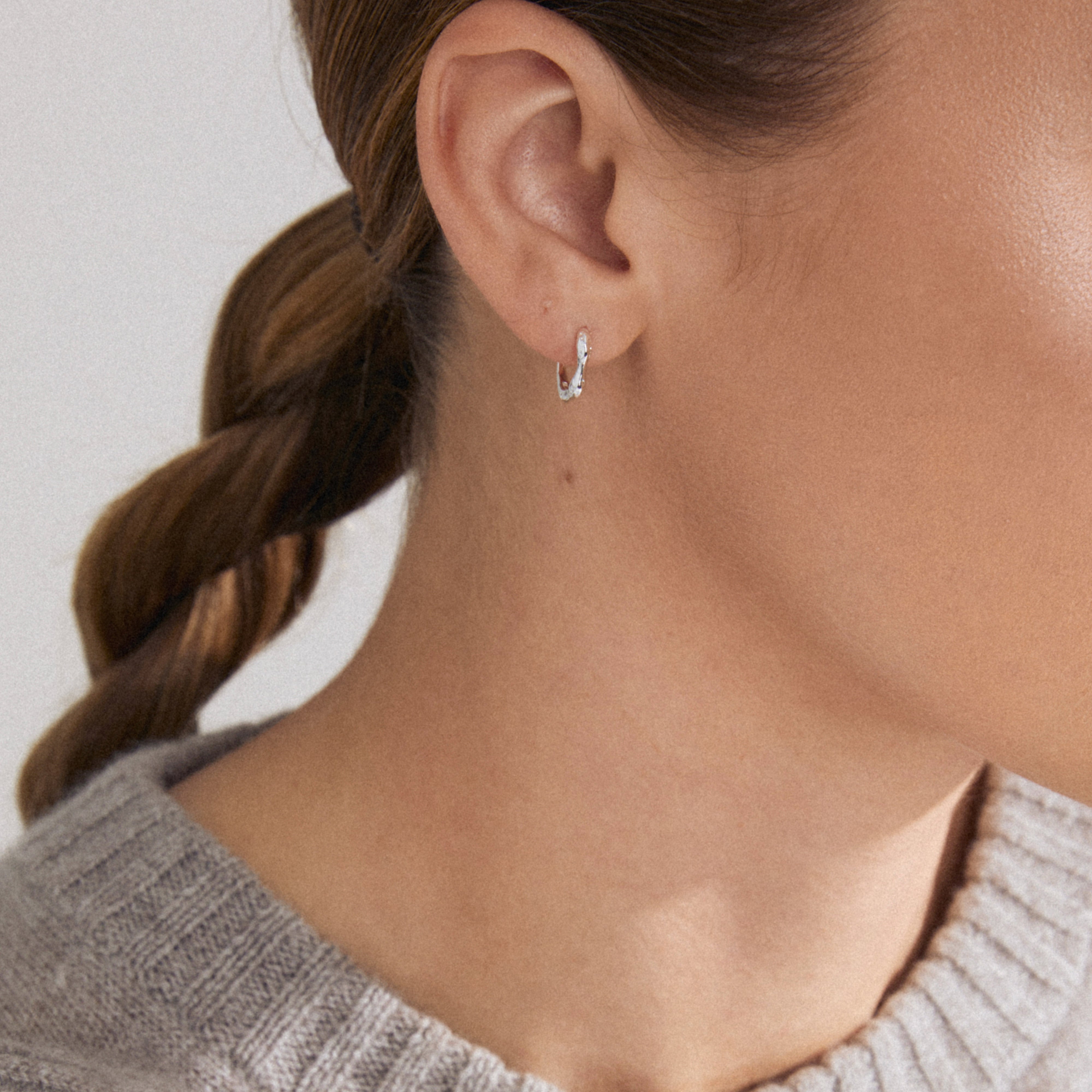 KYRA single earring silver-plated, showcasing a small, textured silver hoop with an organic shape on an ear, paired with a light gray rib-knit sweater.