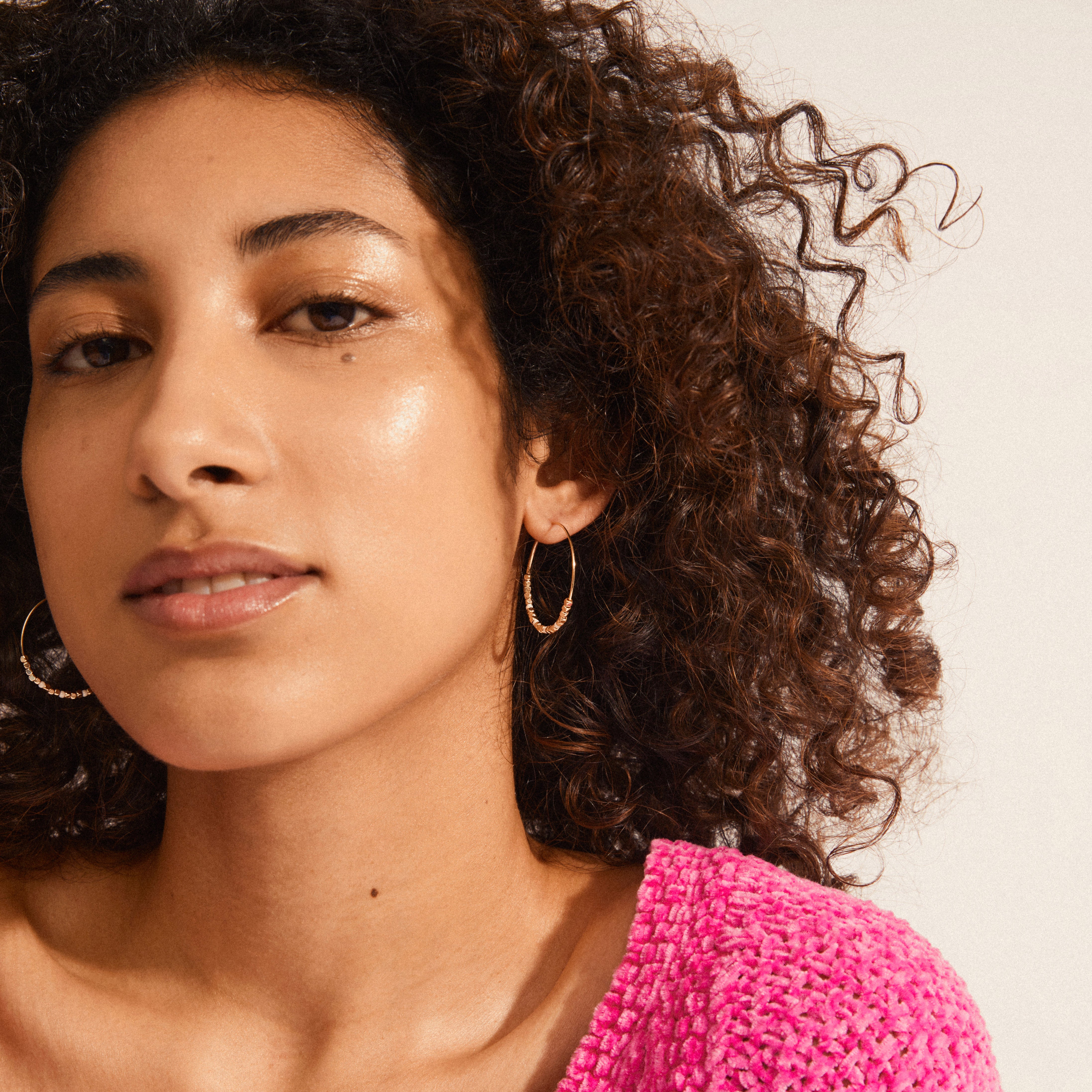 Young woman wearing ROMINA pearl deco hoop earrings, gold-plated, featuring beaded details. Her textured hot-pink top complements the earrings' stylish, feminine design.