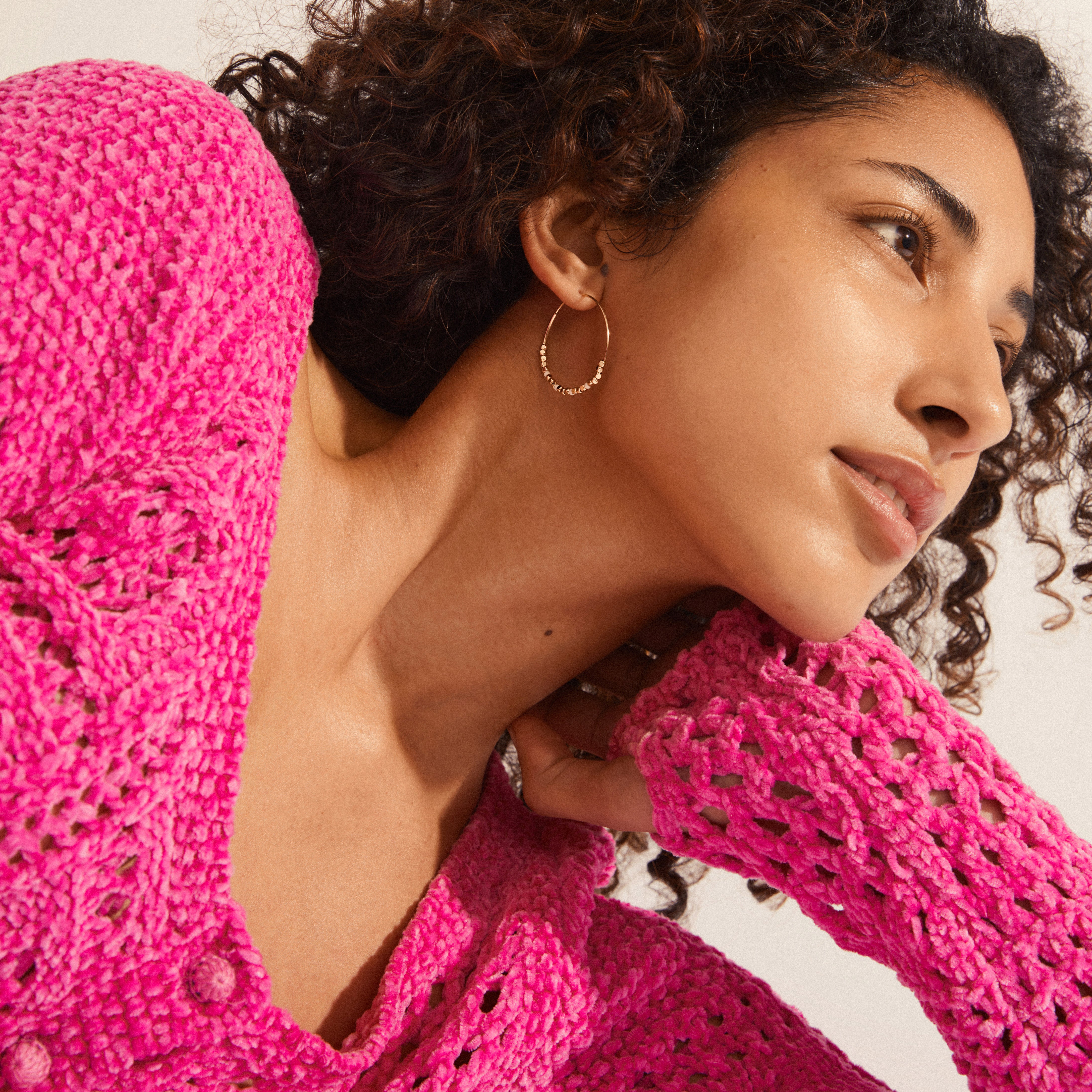 Young woman wears Pilgrim's ROMINA gold-plated hoop earring, featuring metallic beads, paired with a bright pink crocheted sweater, embodying a stylish, feminine summer look.