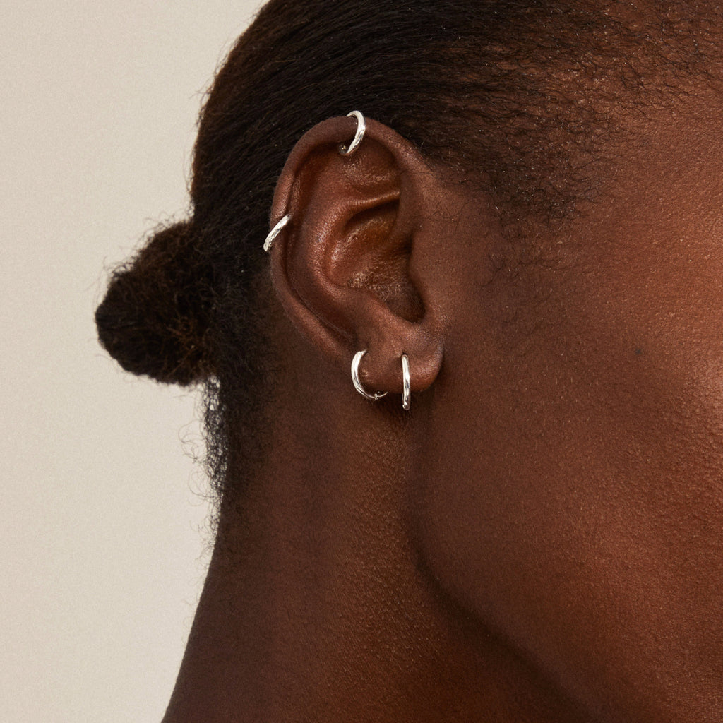Dark-skinned person wears the LEAF 2-in-1 set huggie hoops silver-plated from Pilgrim, featuring small silver hoops in the earlobe and cartilage.