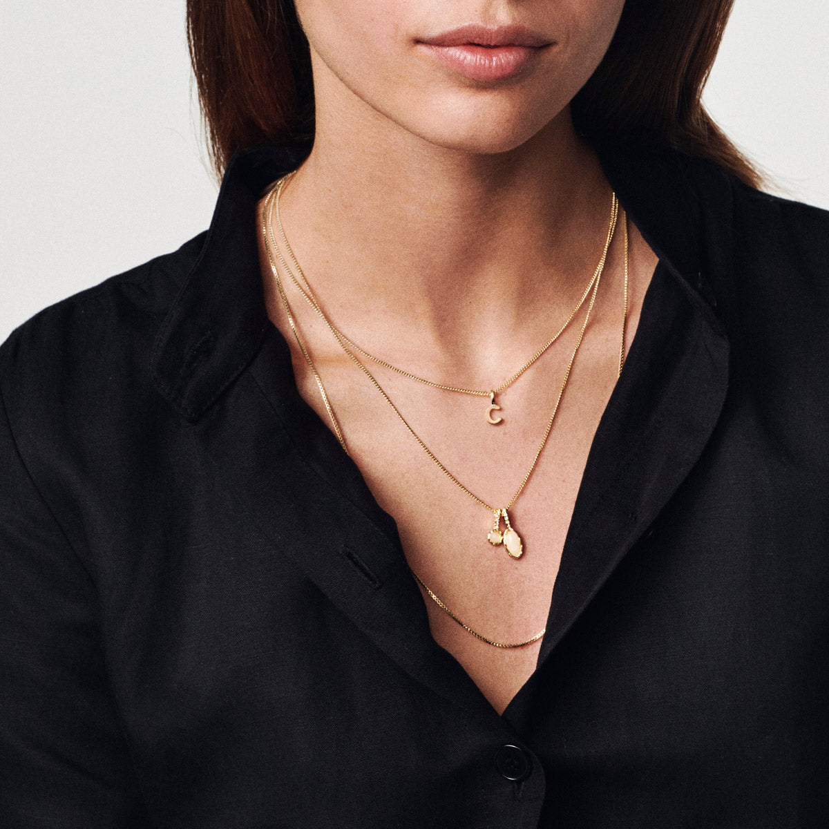 Woman wearing a black top with layered gold chains, featuring the CHARM C pendant, gold-plated, highlighting personalized elegance and Scandinavian style from Pilgrim.