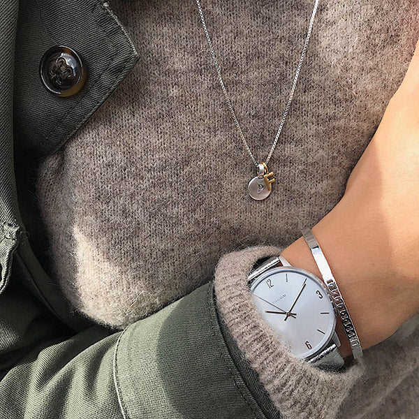 Silver wristwatch and bracelet, featuring a gold-plated CHARM F pendant on a delicate necklace, styled with a knit sweater and olive jacket.