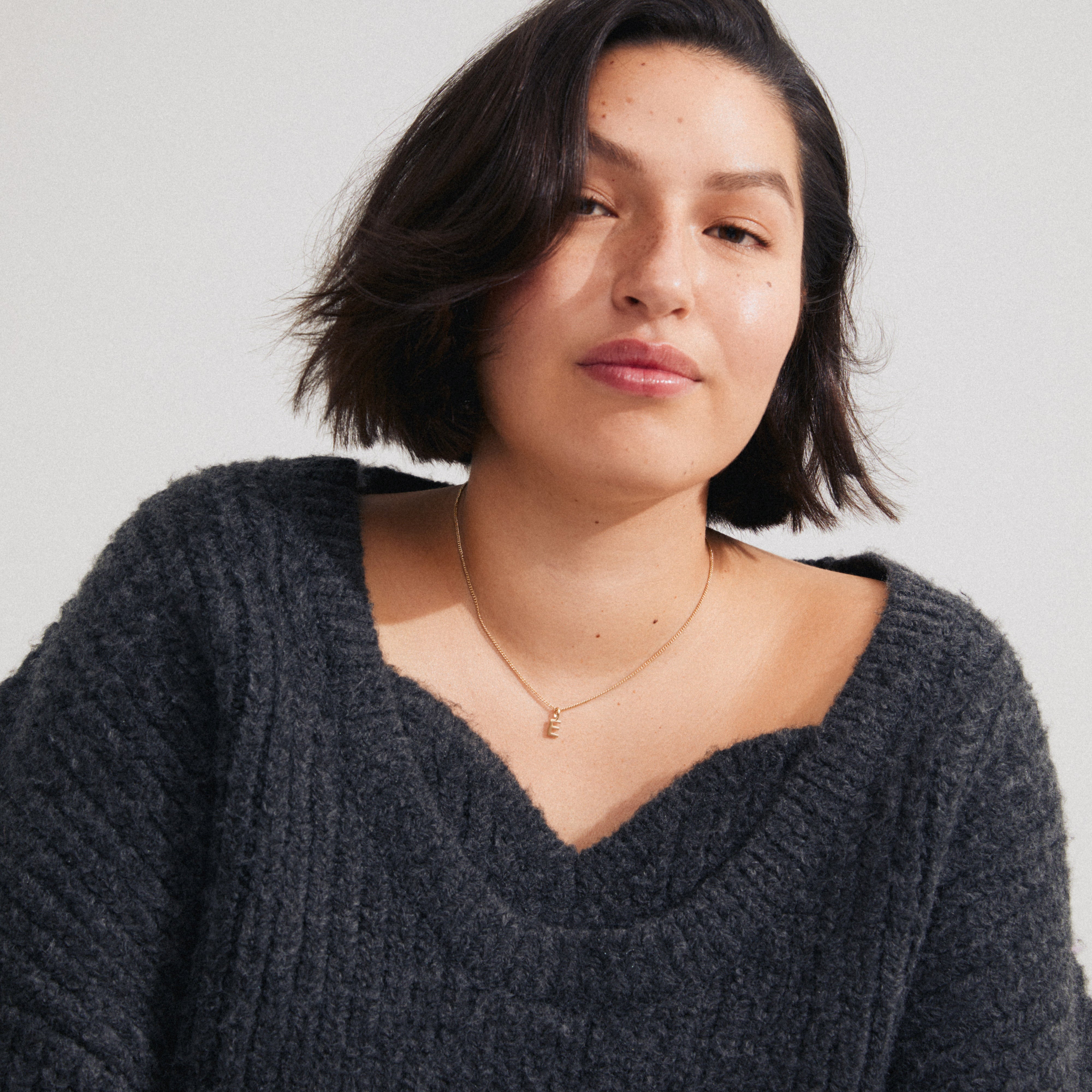 Close-up of a person wearing a CHARM necklace with an E pendant, gold-plated, paired with a chunky-knit charcoal sweater, embodying personalized elegance.