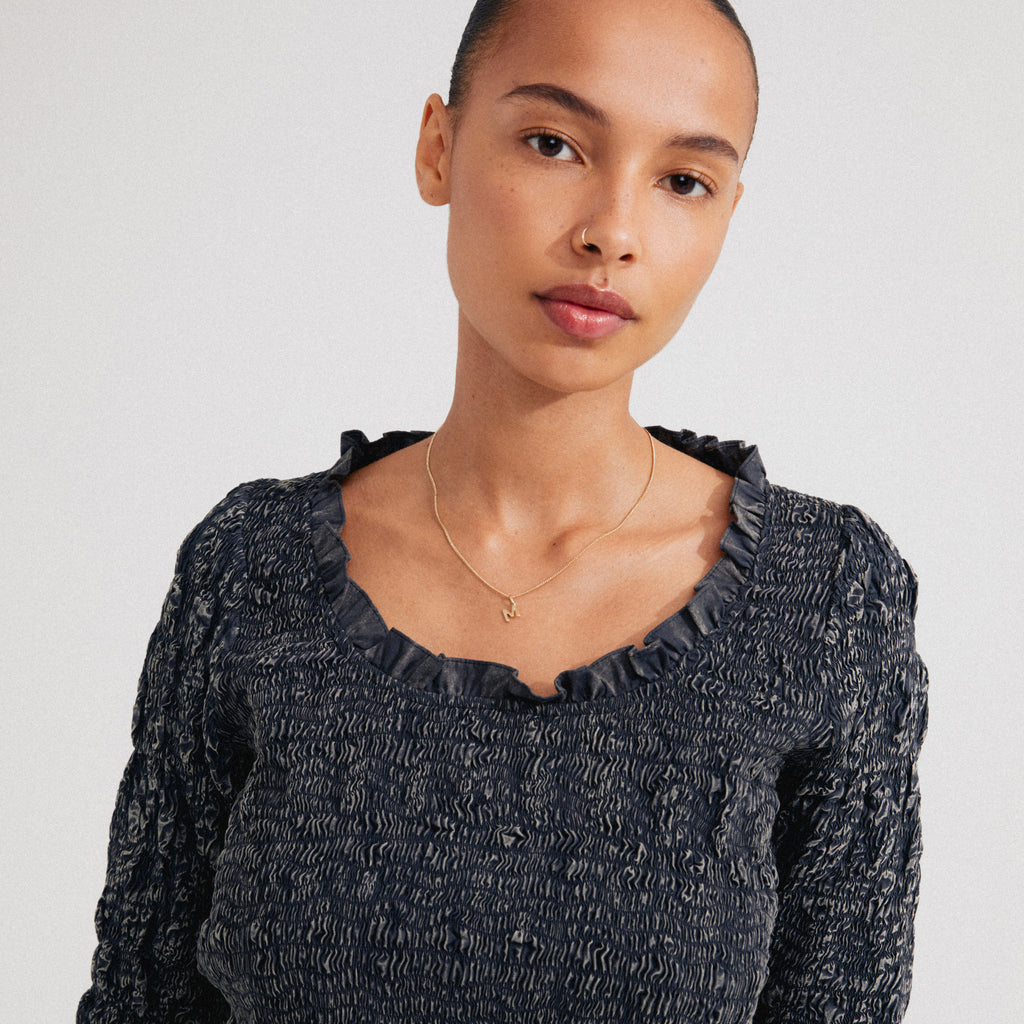 Young woman wears a CHARM necklace with an “M” pendant, gold-plated, complementing her smocked top. The necklace adds a personalized touch to her style.