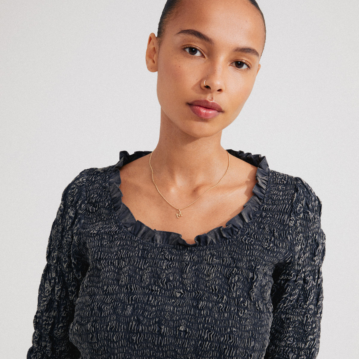 Young woman wears a CHARM necklace with an “M” pendant, gold-plated, complementing her smocked top. The necklace adds a personalized touch to her style.