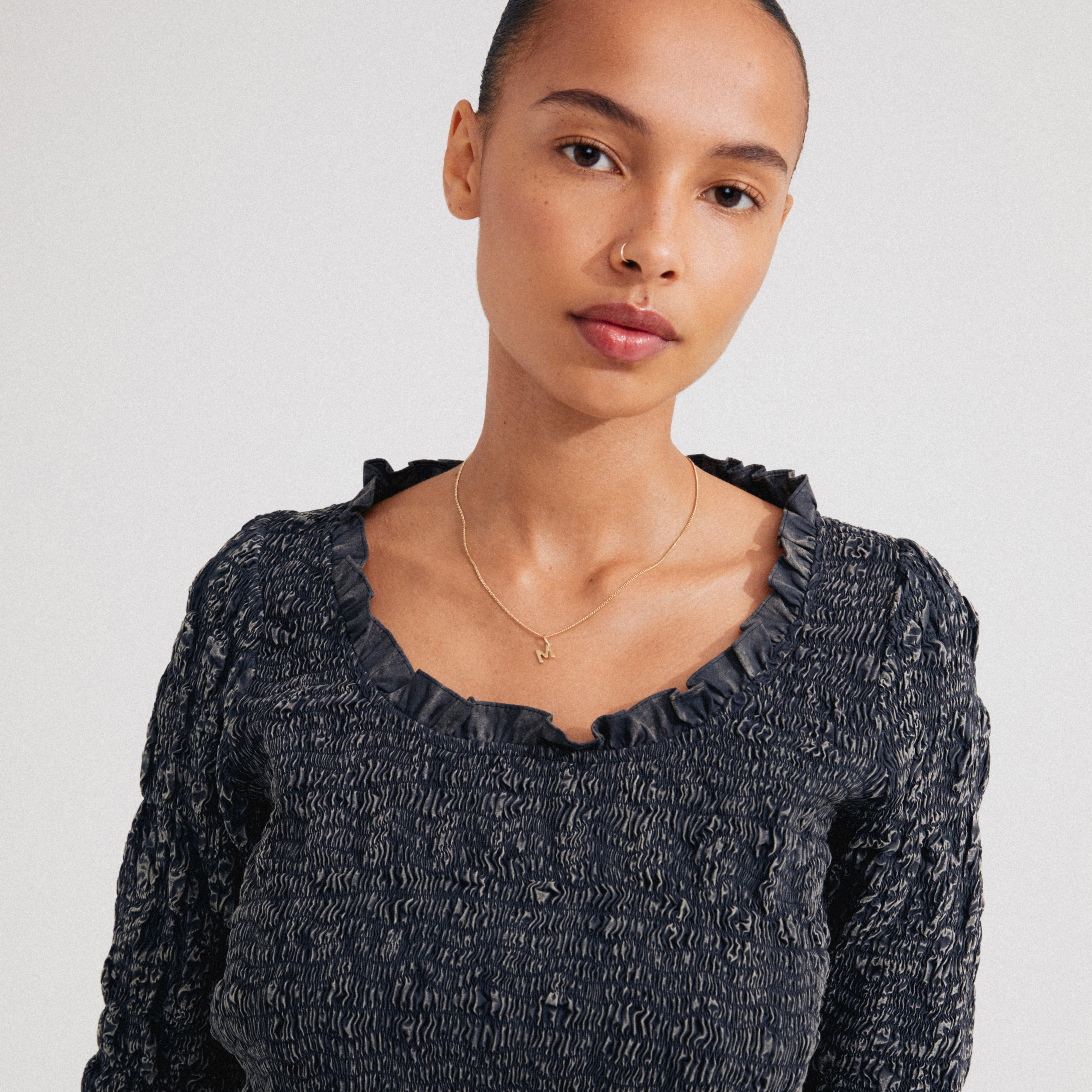 Young woman wears a CHARM necklace with an “M” pendant, gold-plated, complementing her smocked top. The necklace adds a personalized touch to her style.