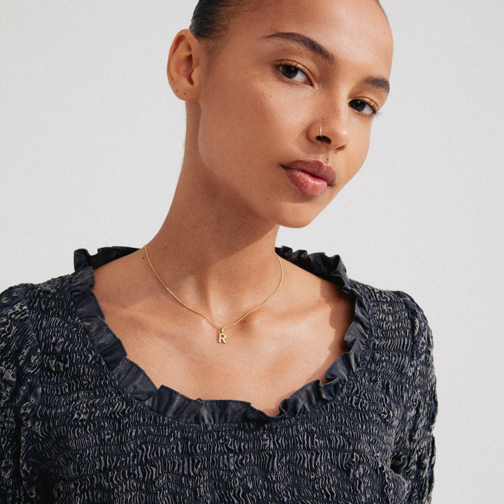 Young woman wearing CHARM necklace with R pendant, gold-plated, featuring a thin chain, showcasing personal style with a minimalist, elegant design.