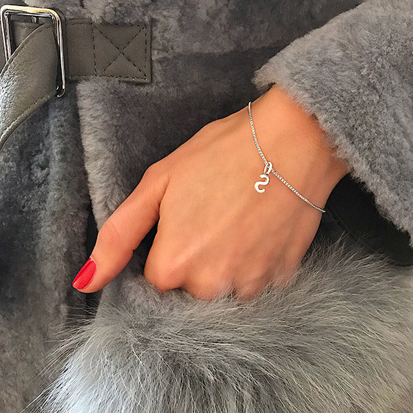 Close-up of a hand in a gray faux-fur coat pocket, showcasing a CHARM S silver-plated pendant on a delicate chain bracelet with a snake-shaped charm.