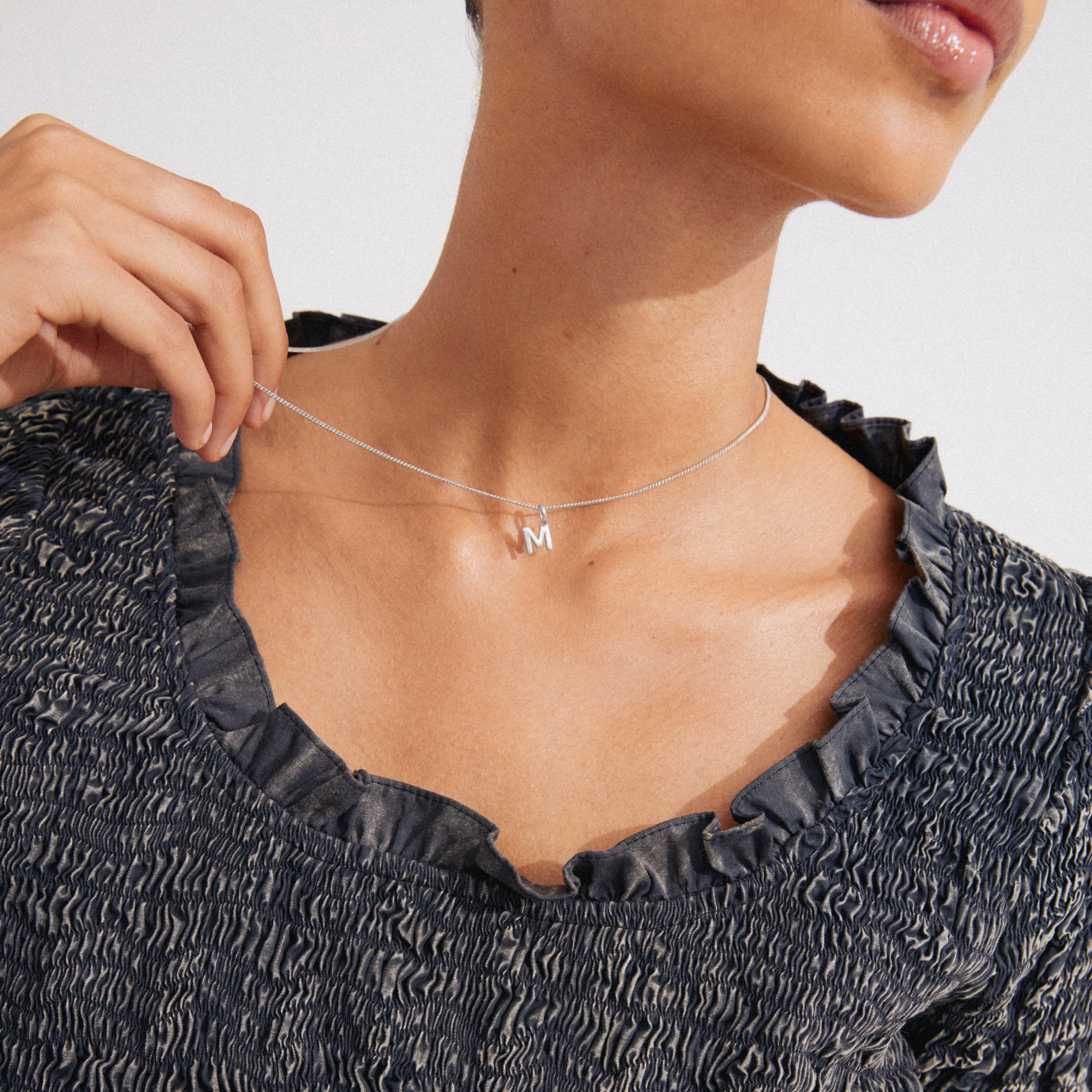Silver CHARM necklace with M pendant, held for visibility, worn with a ruched top. Personalize your style with this 43 cm silver-plated accessory.