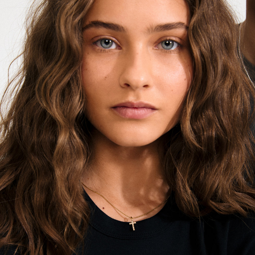 Gold-plated mini cross pendant with sparkling crystals, worn by a woman, symbolizing personal style and statement. Ideal for creating a unique, stacked jewellery look.