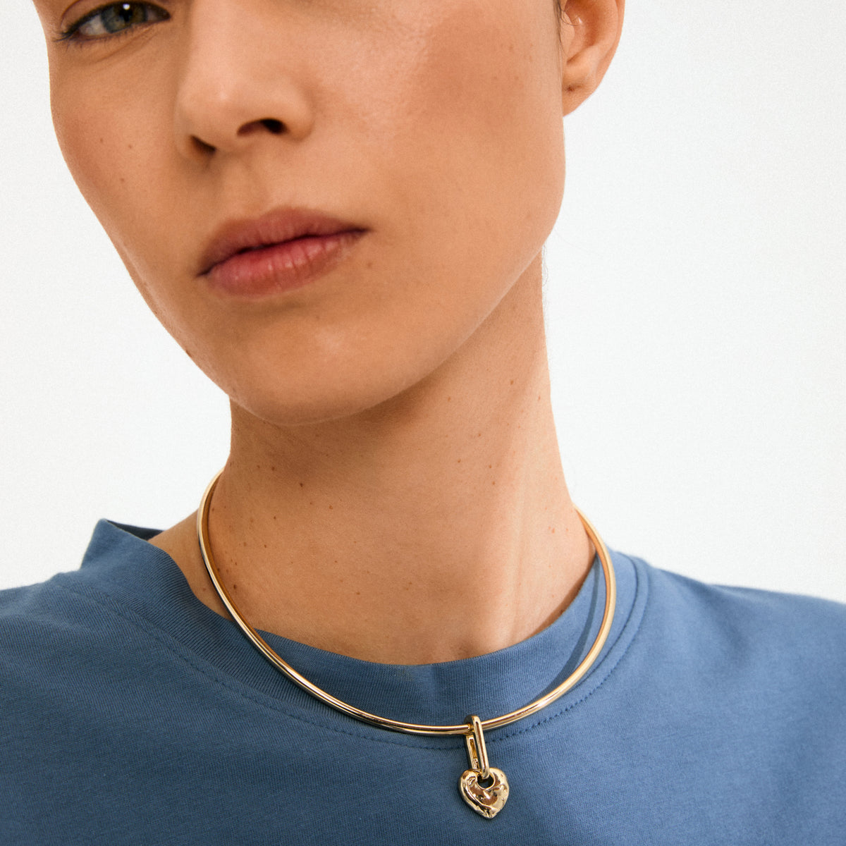 Person wearing a simple blue top with Pilgrim's CHARM rough heart pendant, a unique gold-plated choker-style necklace featuring a rustic heart-shaped padlock.