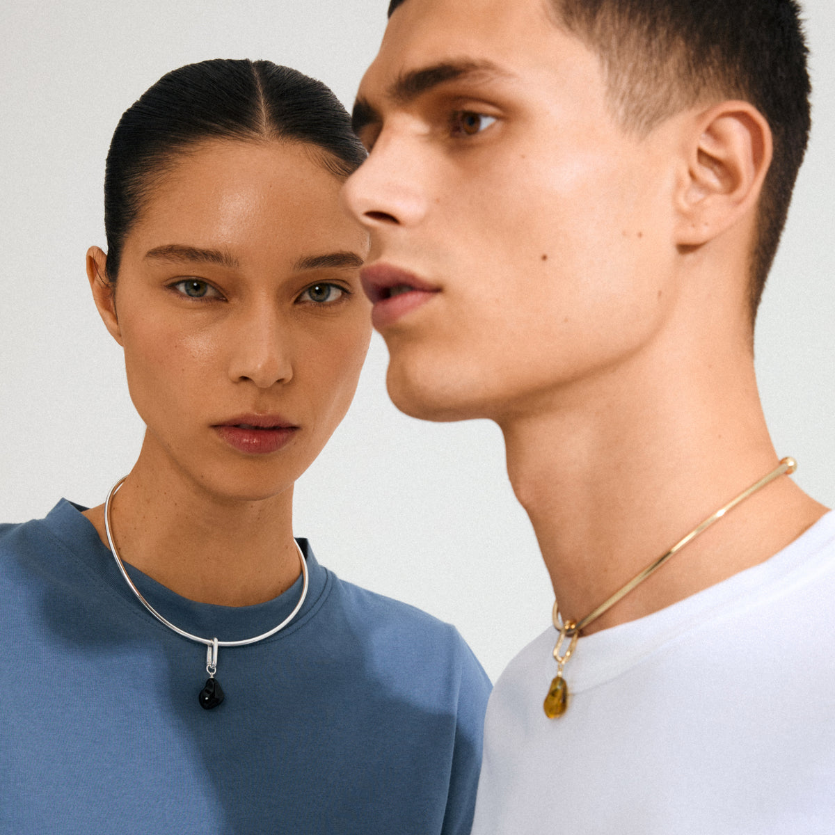 Models showcase Pilgrim's CHARM mold pendant necklaces; woman in silver with black stone, man in gold with amber stone, embodying minimalist elegance and eco-friendly design.