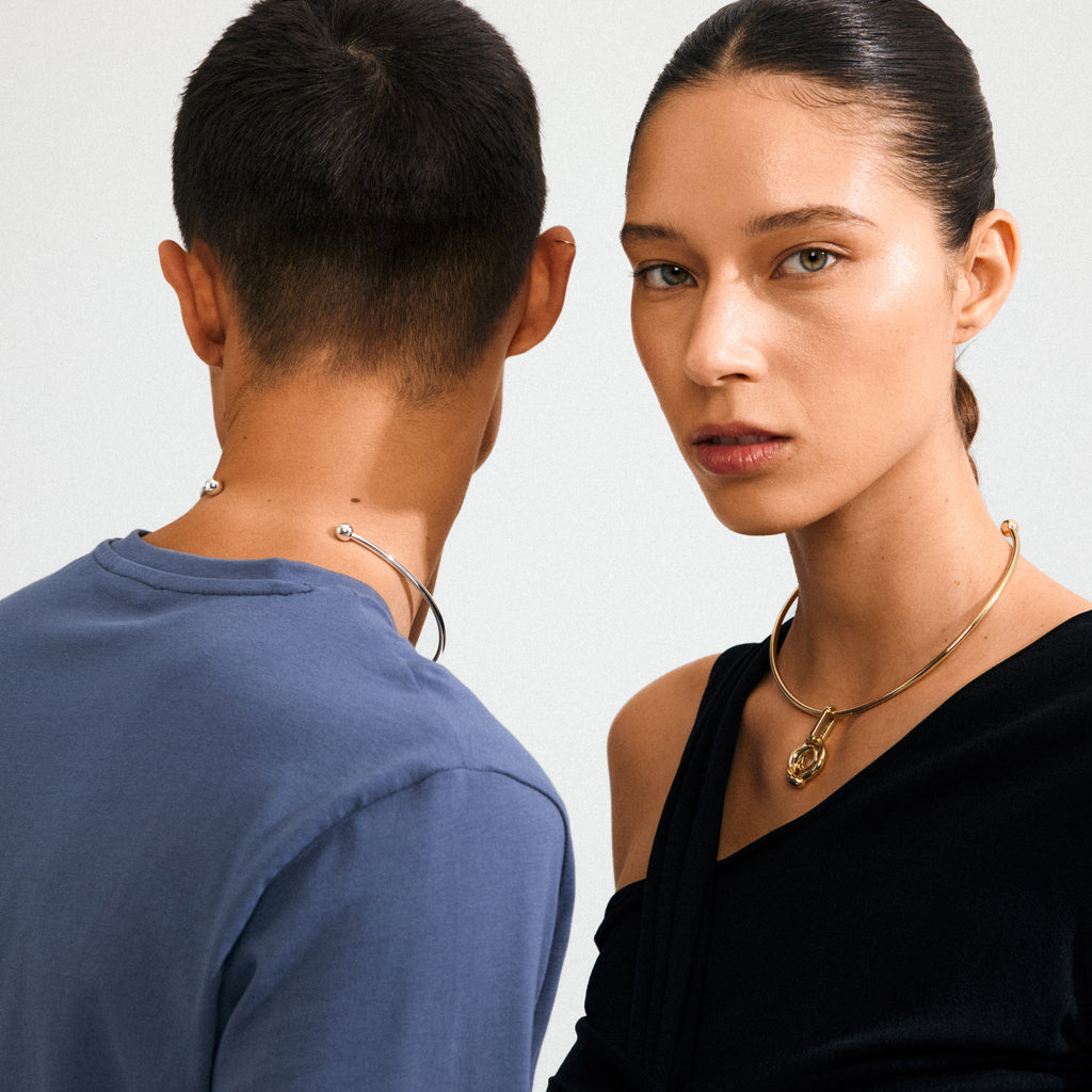 Two models wearing minimalist collars; woman with a gold choker and padlock pendant, man with a silver open-ended collar. Emphasizes Pilgrim's CHARM curl pendant, silver-plated.