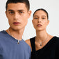 Two models wear Pilgrim's CHARM medallion pendant, silver-plated with a cabochon-cut Tiger Eye stone, showcasing minimalist and stylish jewelry against a plain background.