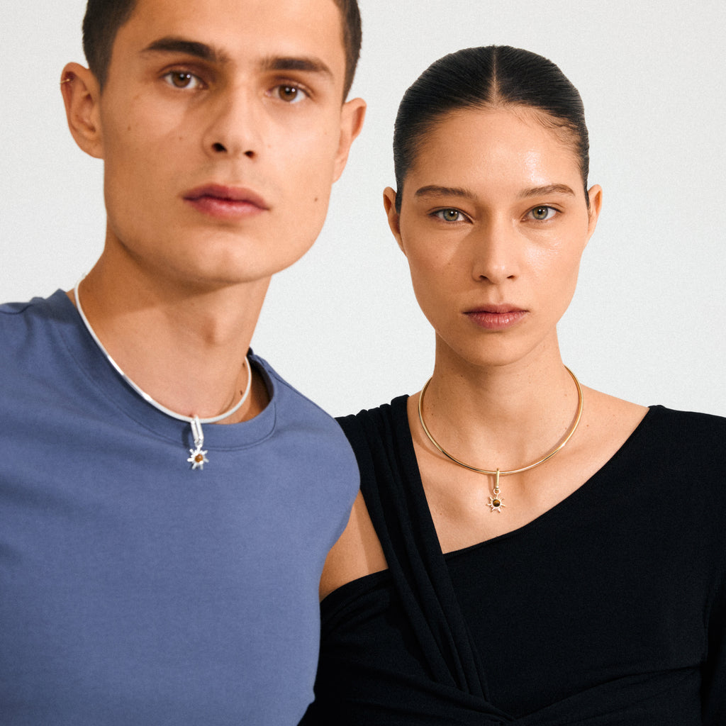 Models showcasing Pilgrim's CHARM bright pendant: male in sleeveless top with silver-plated sun pendant, female in off-shoulder top with gold version, highlighting sleek, minimal design.