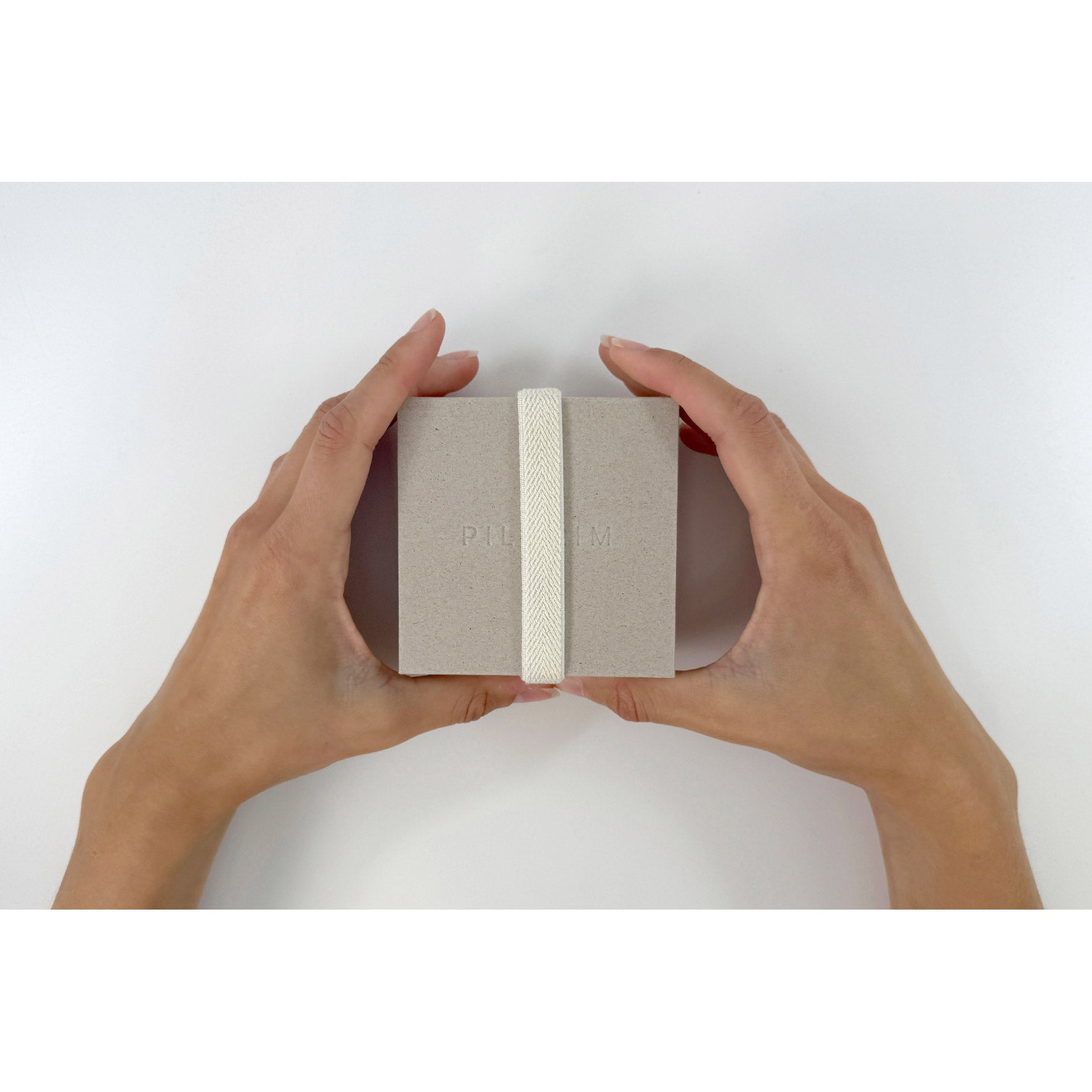 Hands holding a medium-sized Gift box, W85 x H85 x D41 mm, in muted gray with an off-white woven band and debossed PILGRIM logo.