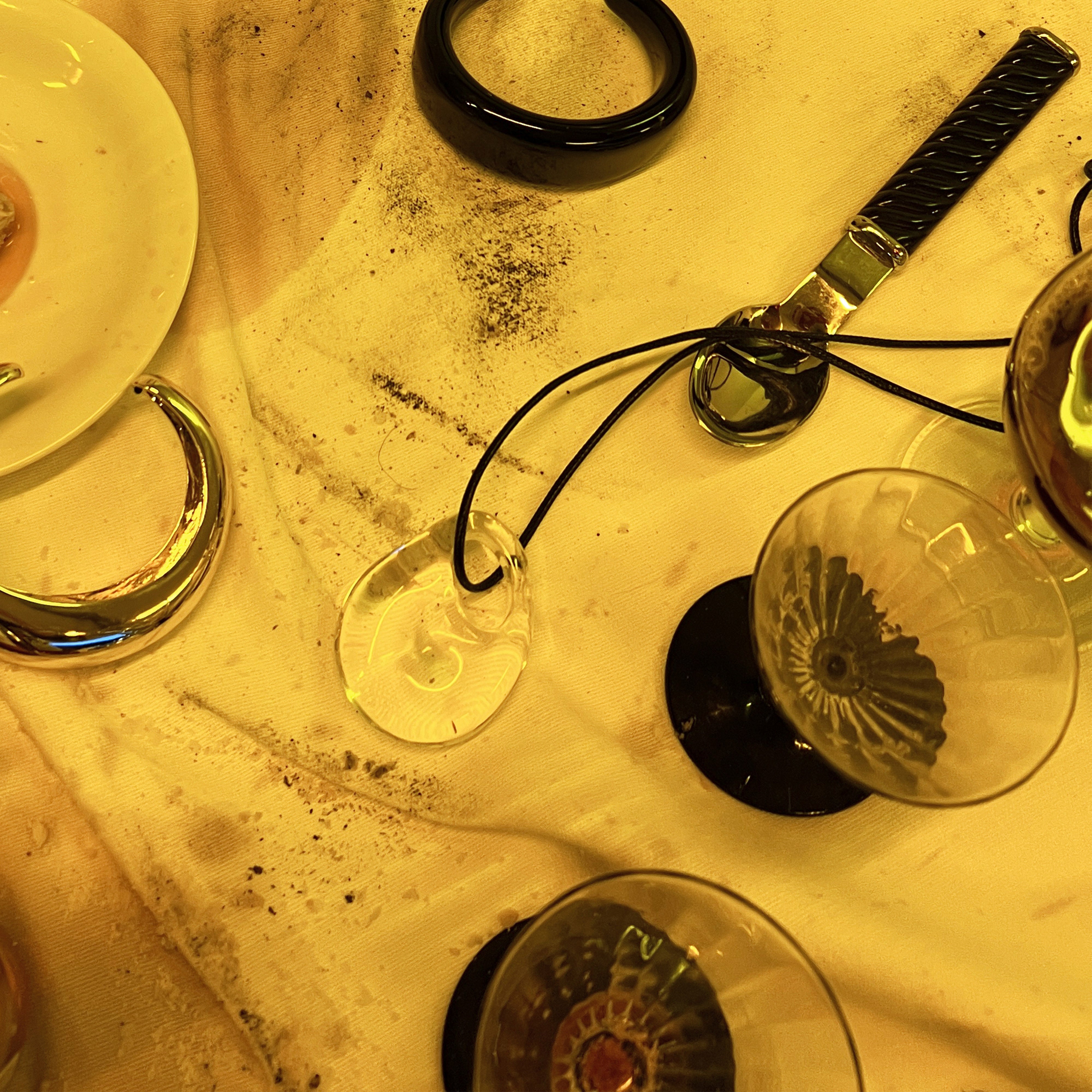 FREJA WEWER x PILGRIM Aqua necklace with a crystal pendant on a black cord, surrounded by jewelry and glassware on a stained tablecloth.