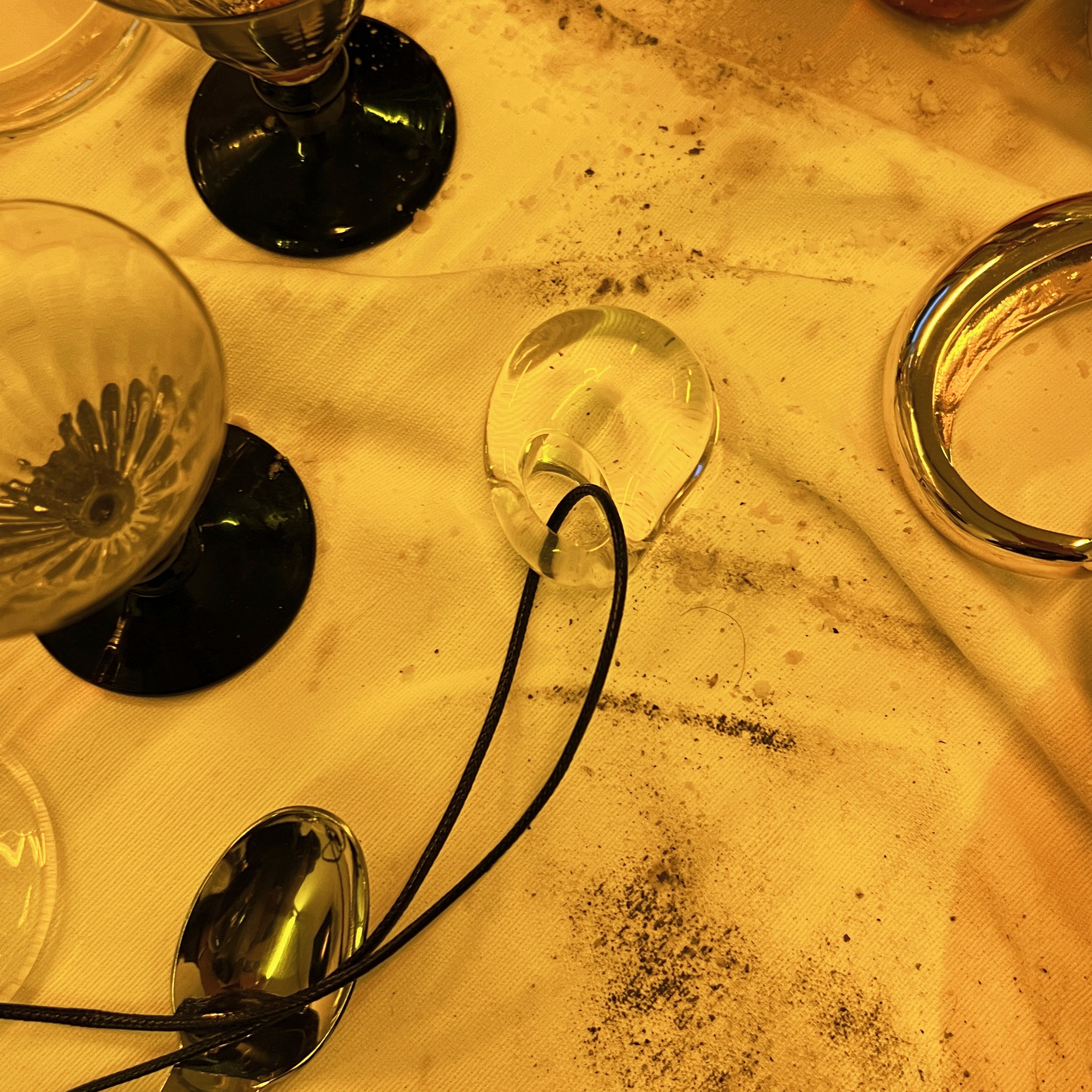 FREJA WEWER x PILGRIM Aqua necklace with clear oval pendant on black cord, surrounded by glass goblets, metal spoons, gold bangle, and dark granules on table.