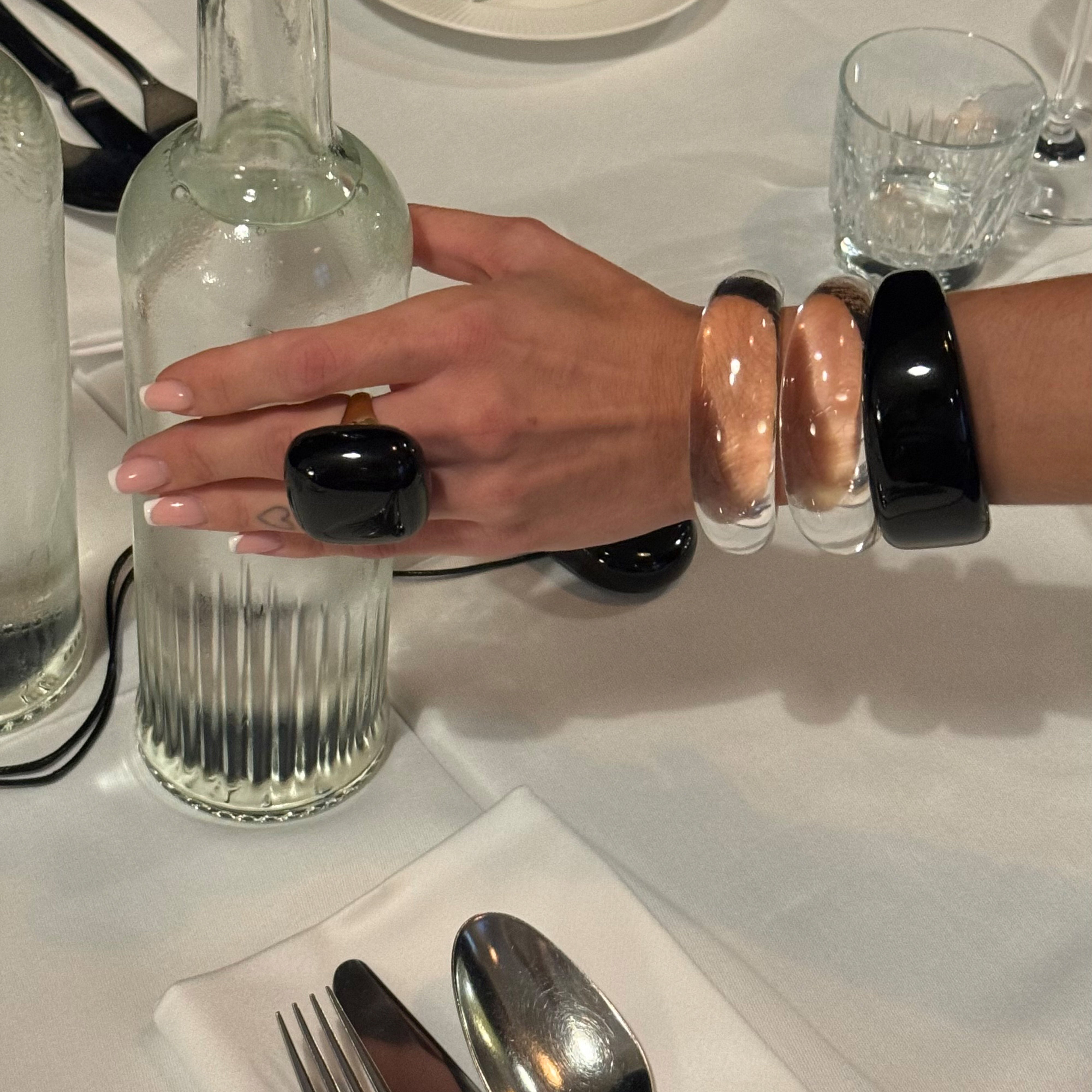 Hand with chunky bracelets, including the FREJA WEWER x PILGRIM Aqua bracelet, crystal, resting on a ribbed glass carafe on a restaurant table.