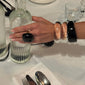 Hand with chunky bracelets, including the FREJA WEWER x PILGRIM Aqua bracelet, crystal, resting on a ribbed glass carafe on a restaurant table.