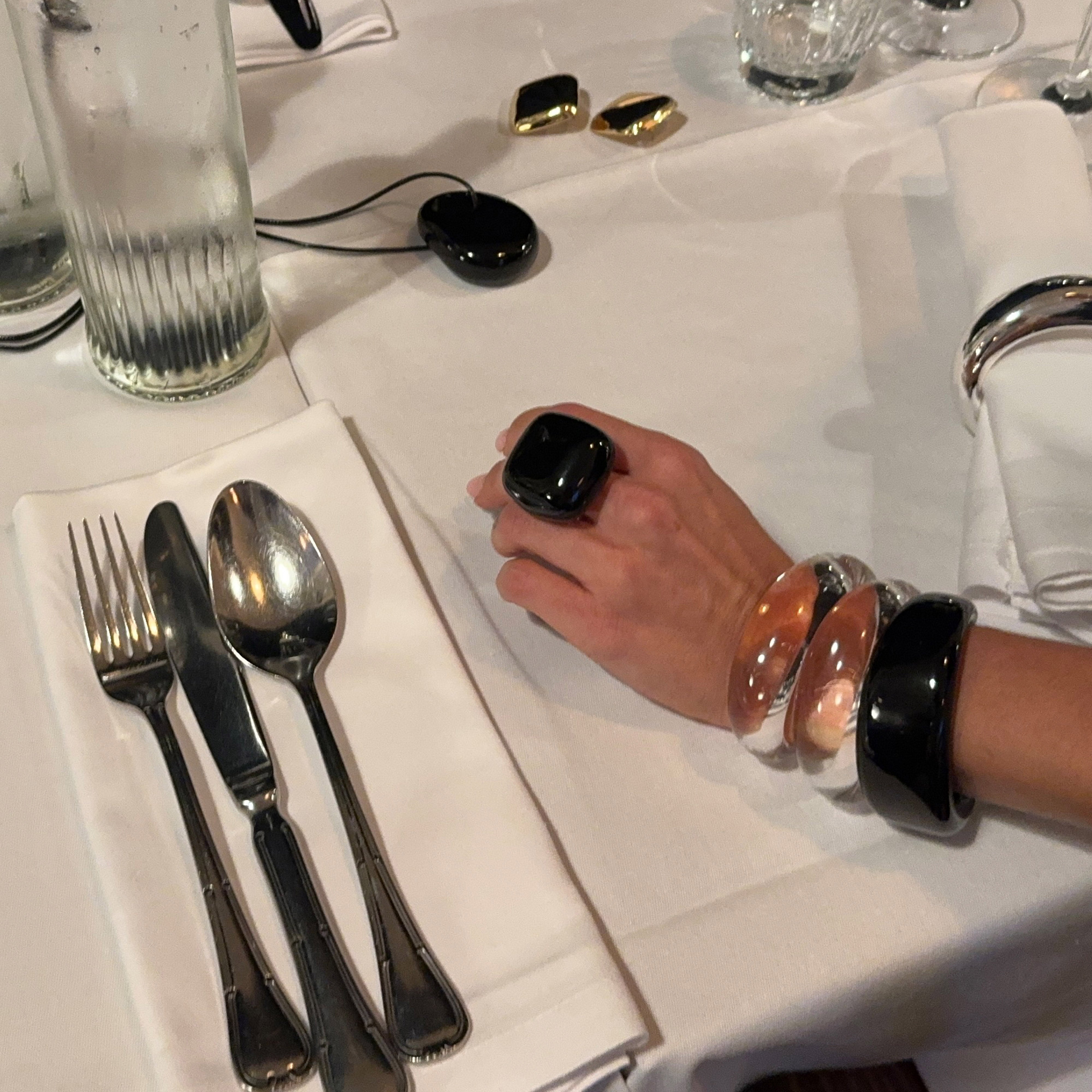 Close-up of a formal place setting showcasing the FREJA WEWER x PILGRIM Aqua bracelet, crystal, worn on a hand with other resin bangles.