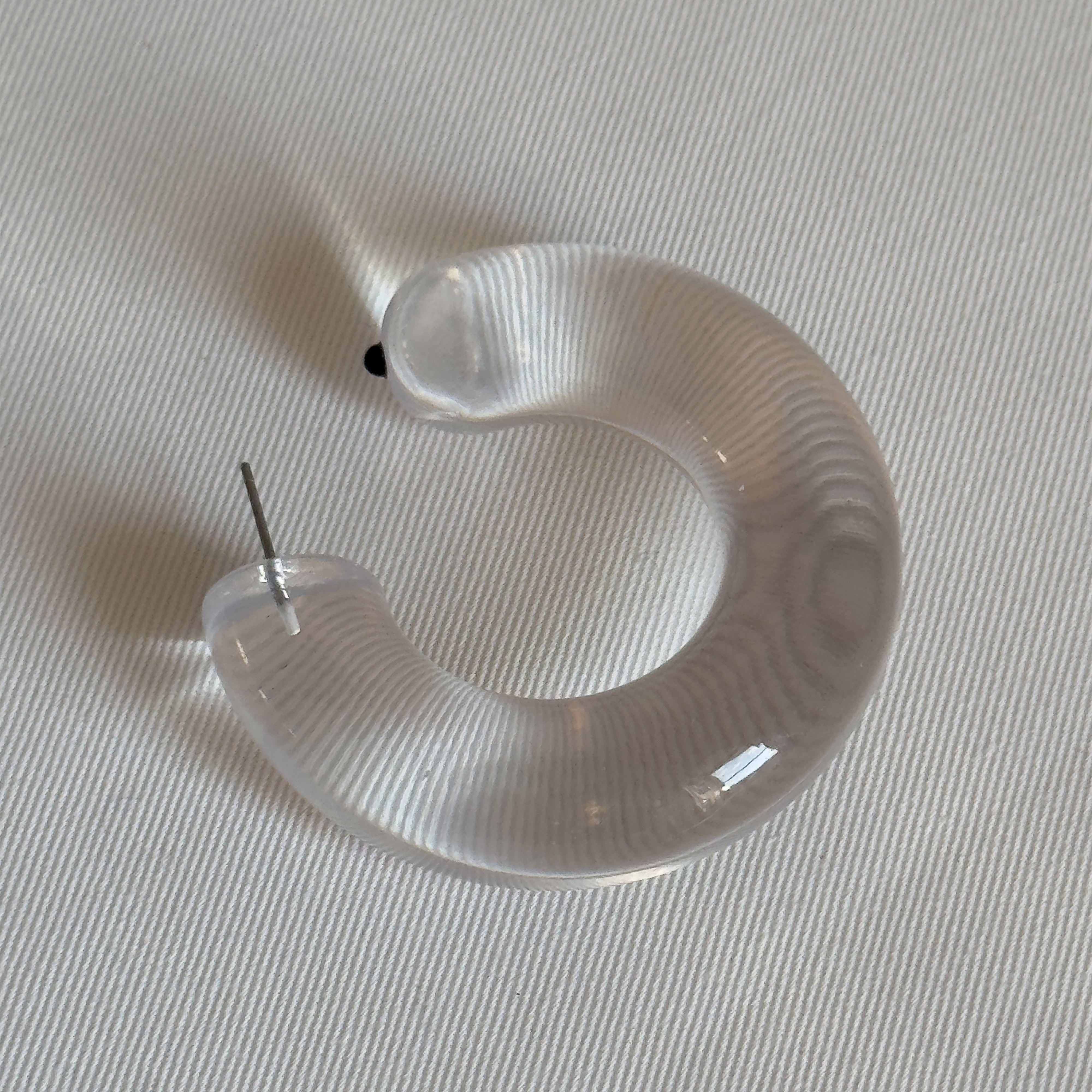 FREJA WEWER x PILGRIM Aqua hoops large, crystal. A single translucent hoop earring, showcasing an oversized, chunky silhouette with a visible metal post on a textured surface.