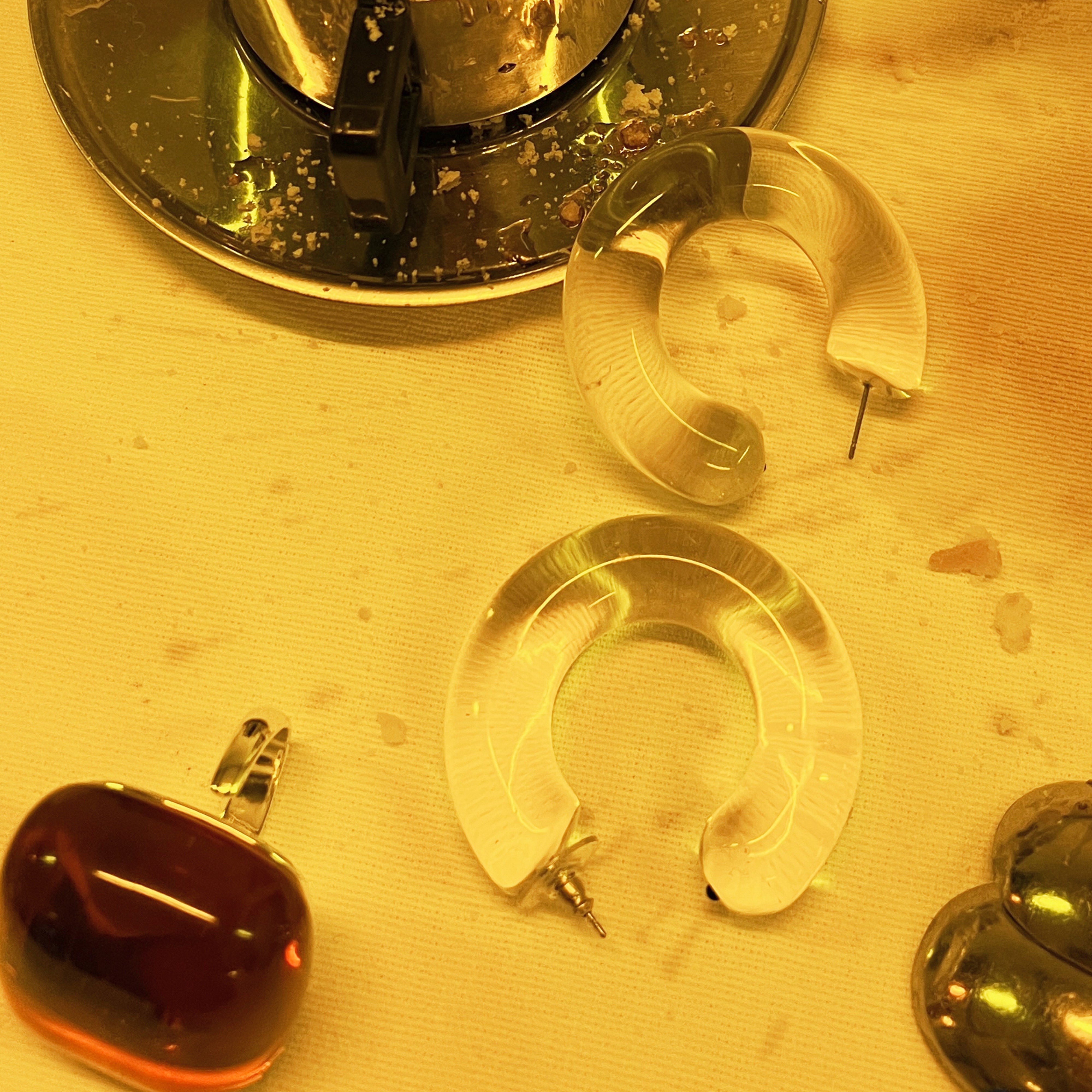 Close-up of FREJA WEWER x PILGRIM Aqua hoops large, crystal. These clear, crescent-shaped Lucite earrings highlight their oversized, trendy design amidst a tabletop setting.