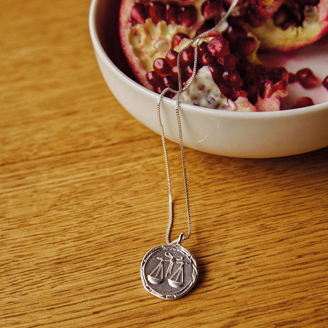 LIBRA Zodiac Sign Coin Necklace, silver-plated, featuring a zodiac motif and constellation pendant, displayed alongside a pomegranate, highlighting its elegant design.