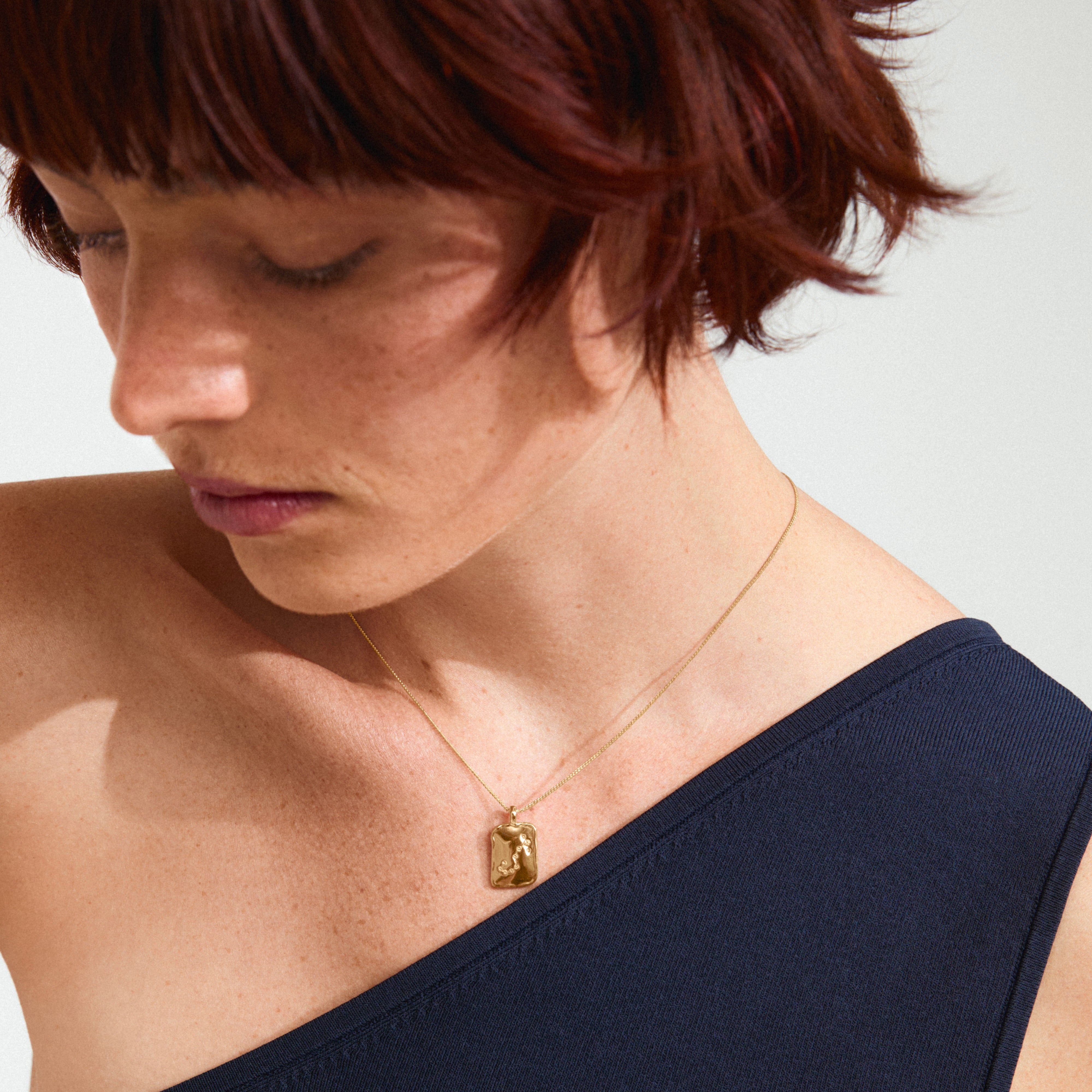 SCORPIO Zodiac Sign necklace, gold-plated, features a square coin pendant with a dual design, worn by a woman with short red hair in a black dress.