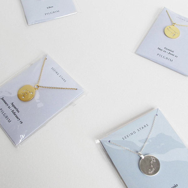 LEO Zodiac Sign Coin Necklace, gold-plated with a Leo star sign pendant adorned with crystal stones, displayed in packaging alongside other necklaces.