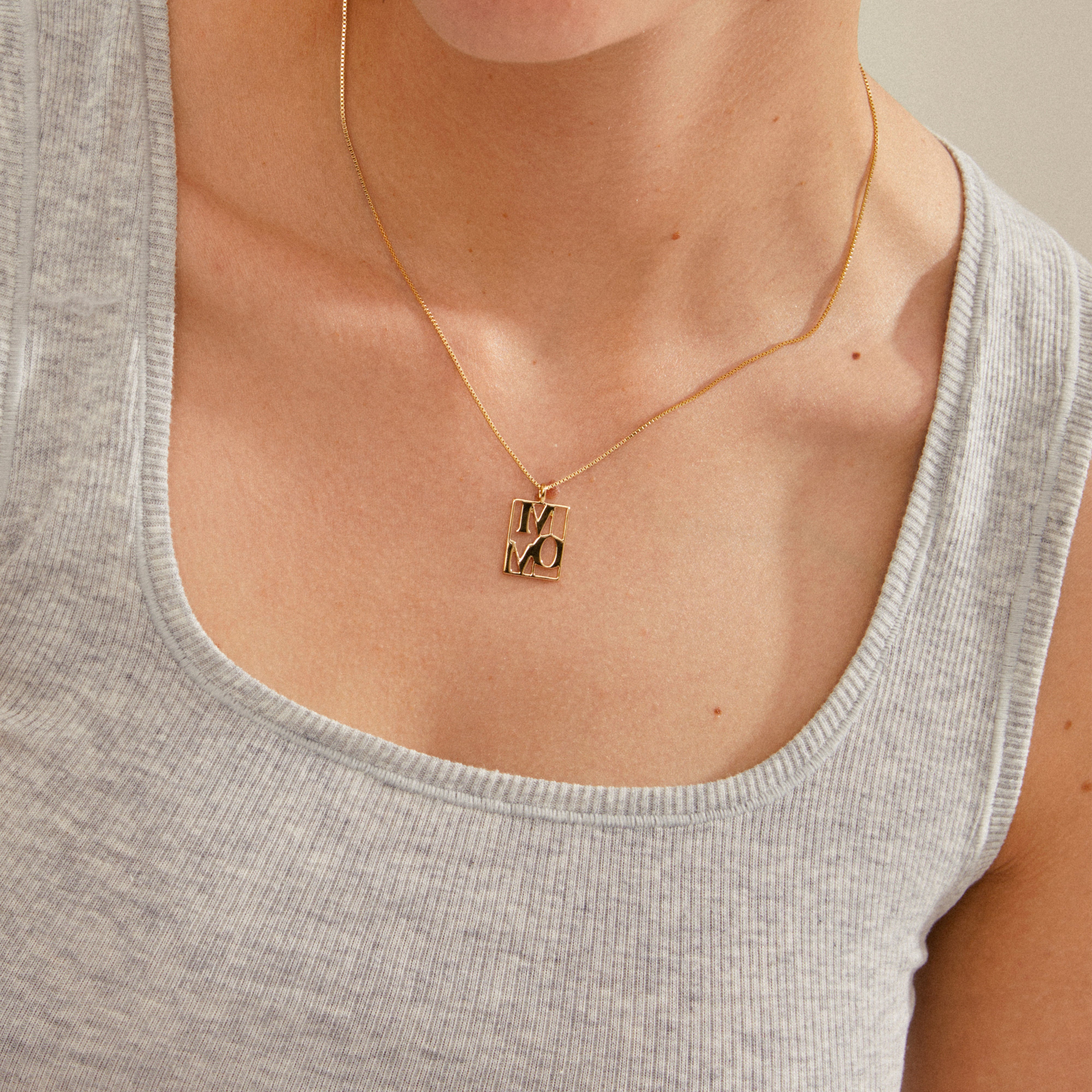 LOVE TAG, MOM necklace gold-plated, featuring a shiny rectangular pendant on a light grey tank top, exemplifying Pilgrim's blend of timeless and trendy Scandinavian design.