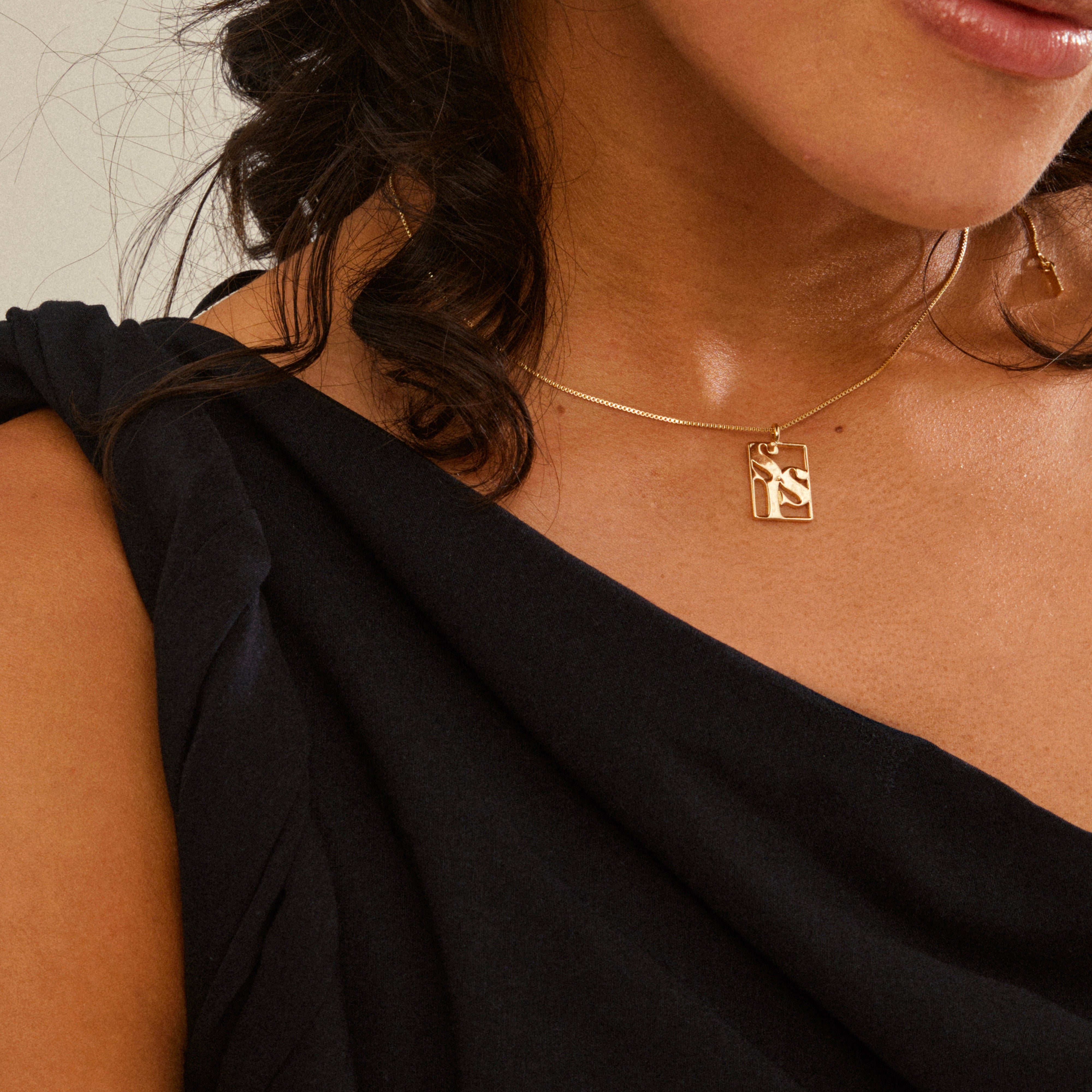 Close-up of a person wearing a LOVE TAG, SIS gold-plated necklace by Pilgrim, featuring a delicate chain and rectangular pendant with cut-out letters.