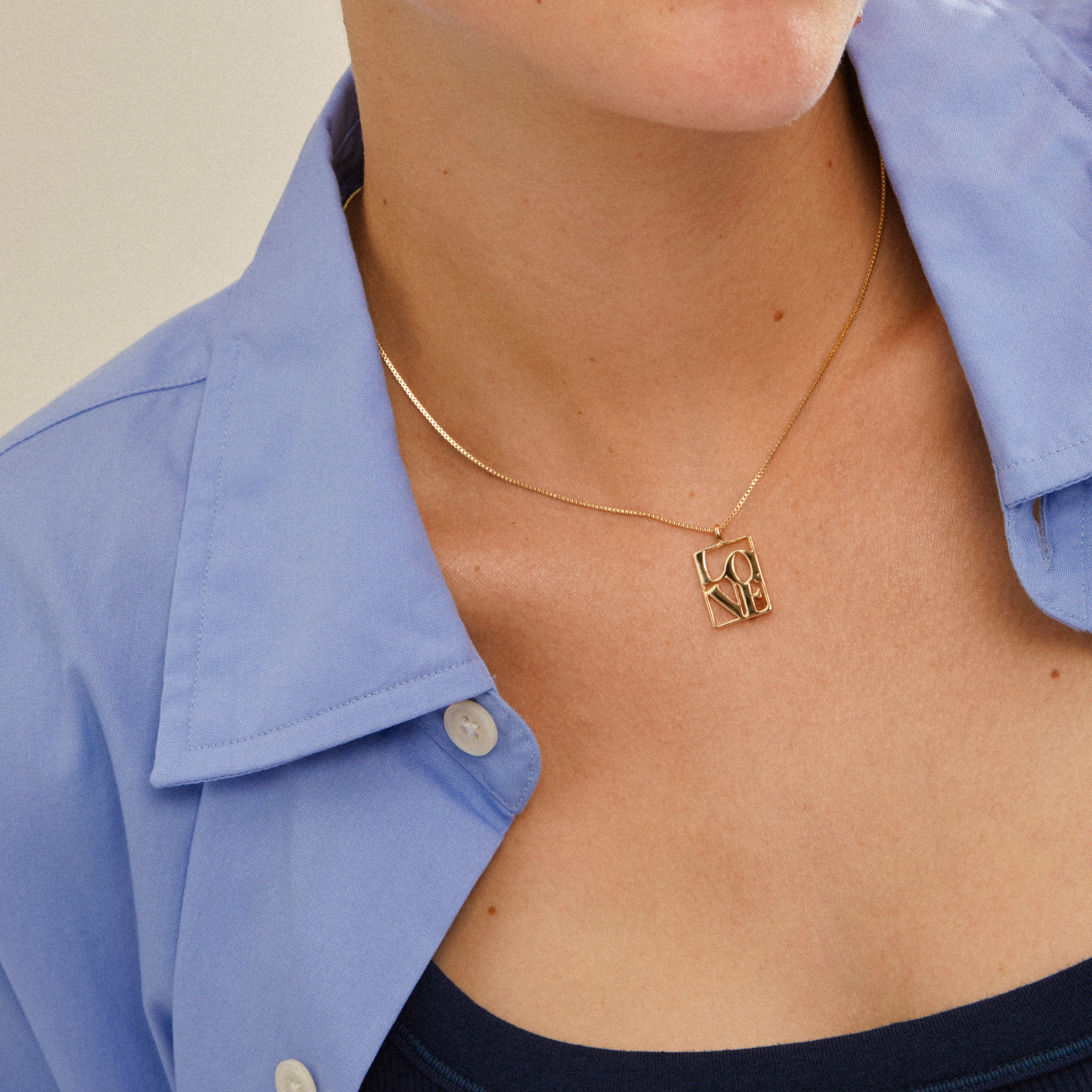 Close-up of a gold-plated LOVE TAG necklace from Pilgrim, featuring a rectangular pendant with LOVE, worn over a light-blue shirt for a layered look.