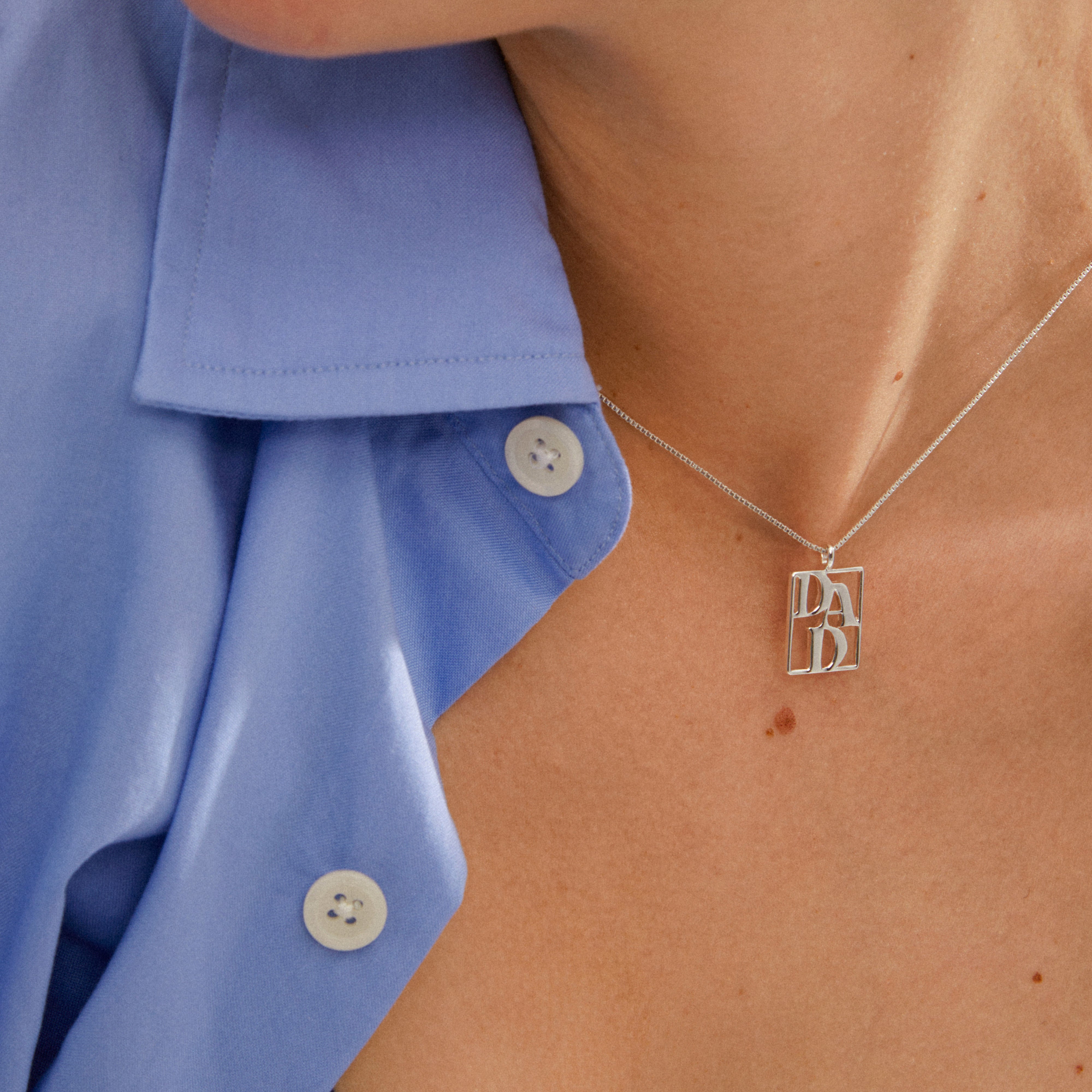 LOVE TAG, DAD necklace silver-plated on a person wearing a light blue shirt, showcasing a rectangular pendant with DAD design, symbolizing love and style.
