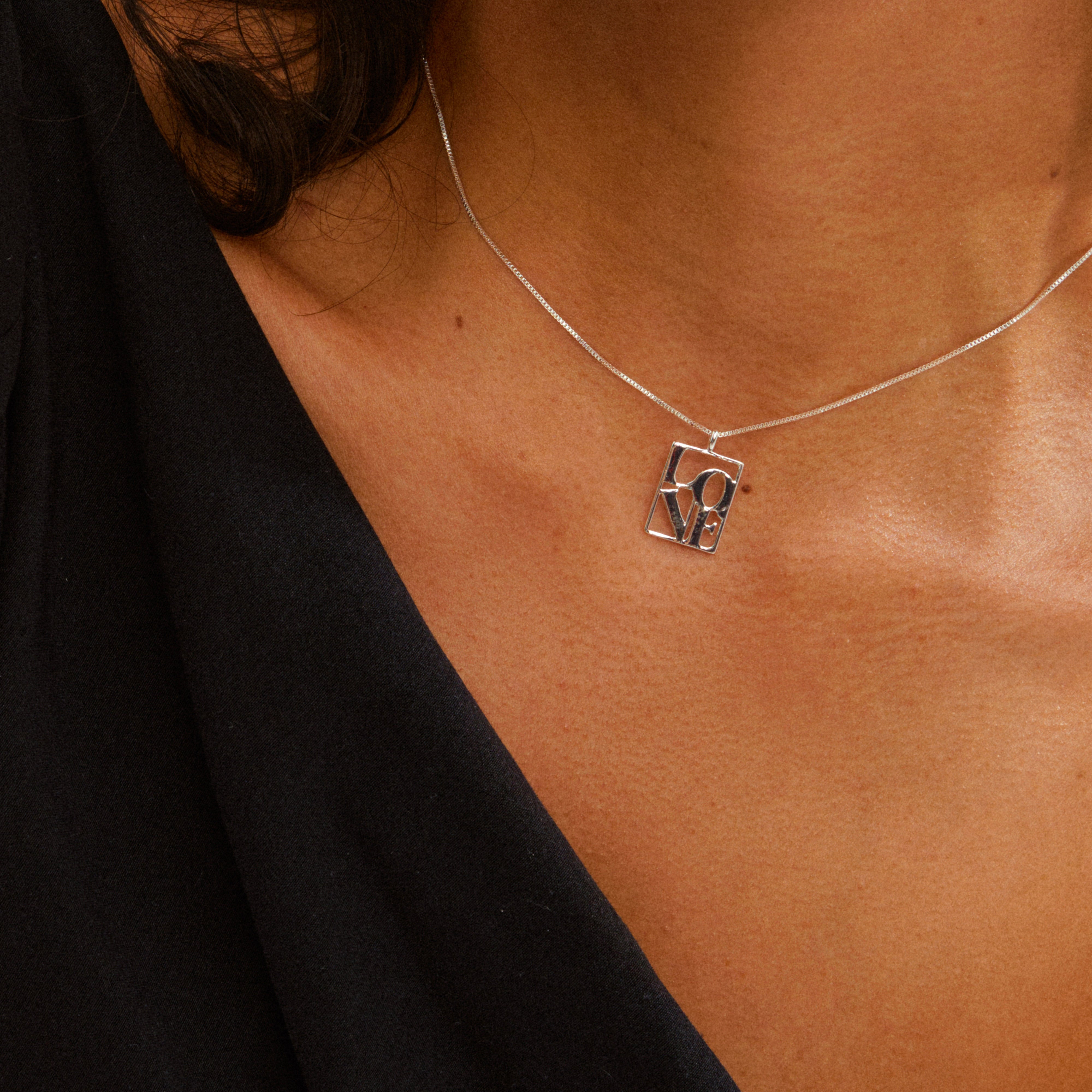 Close-up of a silver-plated LOVE TAG necklace from Pilgrim, featuring a delicate chain and cut-out LOVE pendant, worn with a dark V-neck top.