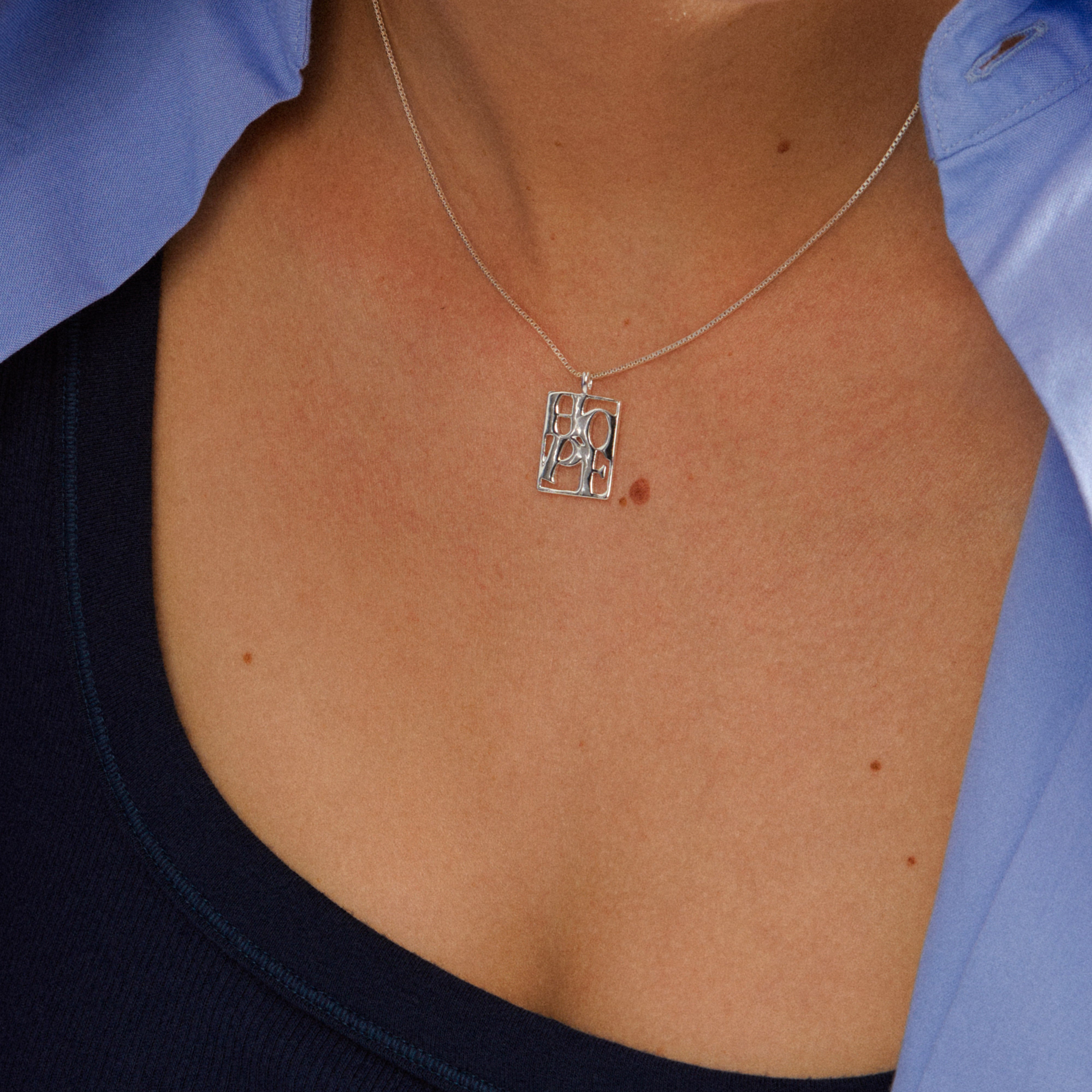 Close-up of a silver-plated LOVE TAG, HOPE necklace by Pilgrim, featuring a small rectangular pendant with DOPE cutouts, worn over a dark blue tank top.