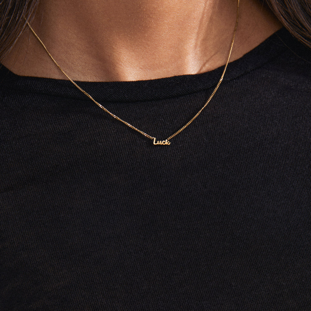 Gold-plated LUCK necklace with a cursive script pendant, worn by a person. A versatile, genderless accessory perfect for layering and personal styling.