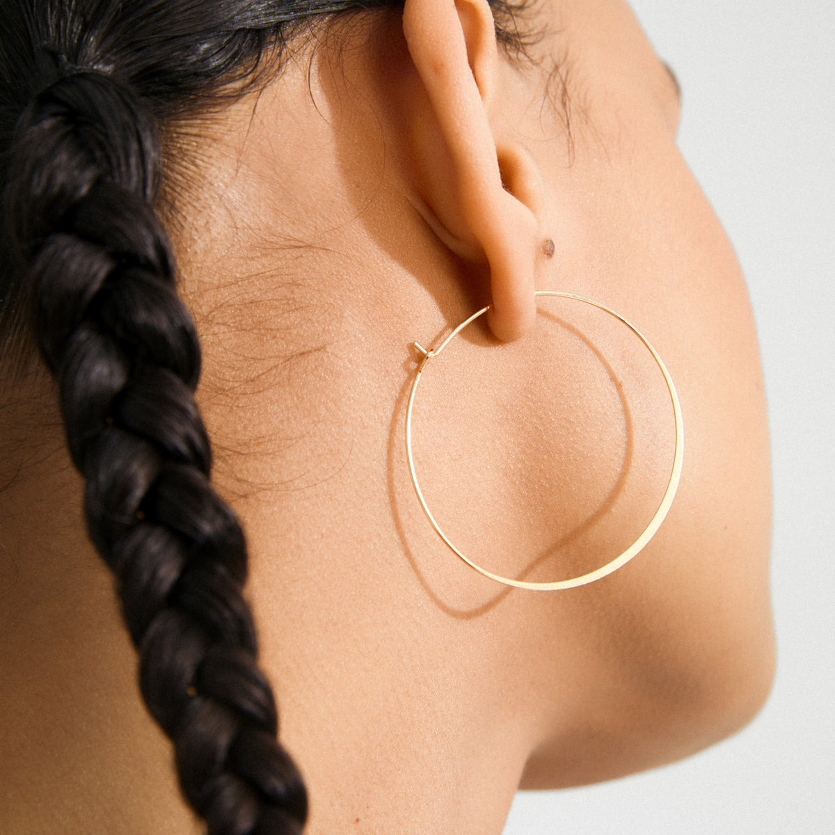 Close-up of TILLY large gold-plated hoop earring adorning an ear with a long dark braid, embodying sleek Scandinavian style from Pilgrim.