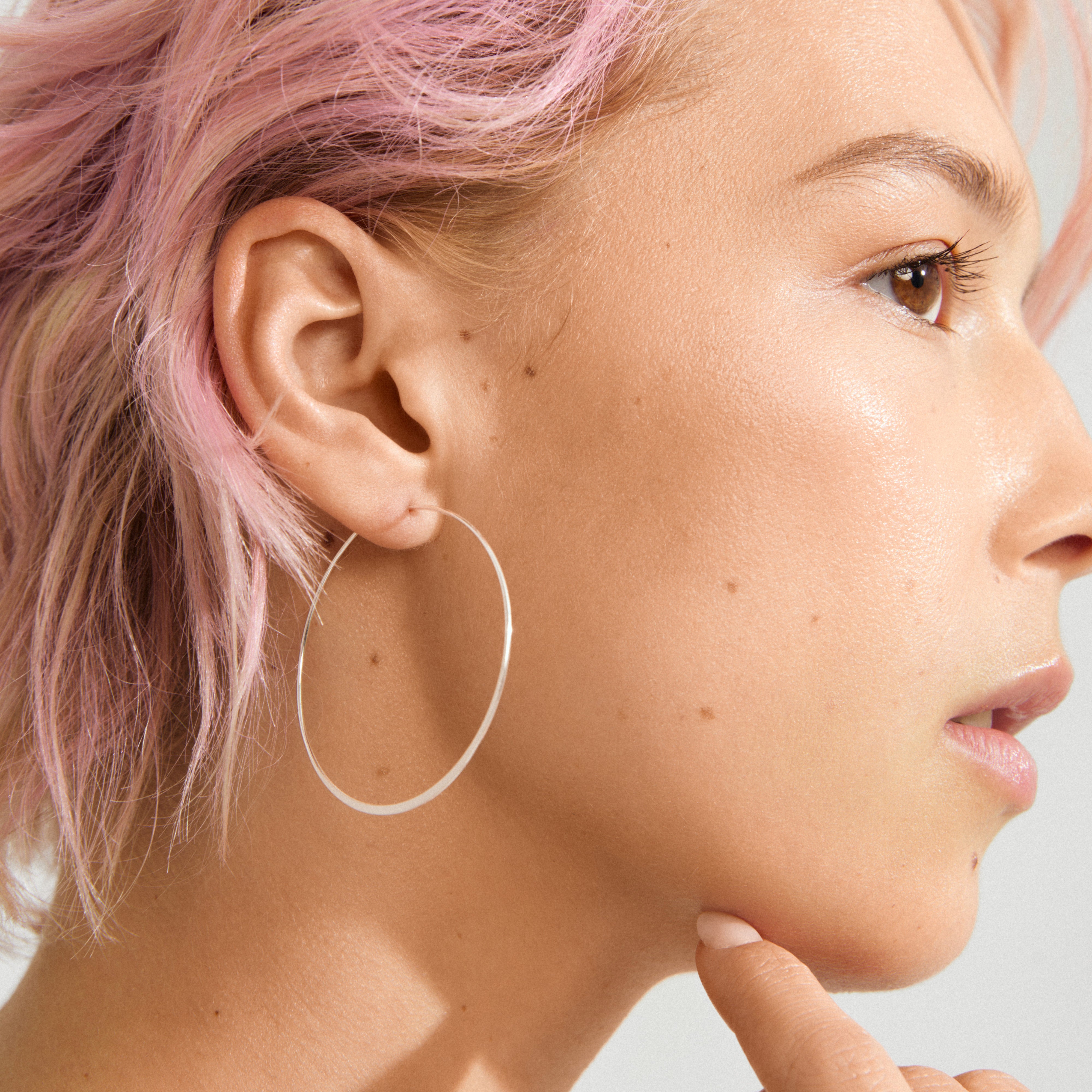 Side profile of a person wearing Pilgrim's TILLY large silver-plated hoop earring, showcasing its sleek, airy design. Hand lightly touching chin, emphasizing minimalist style.