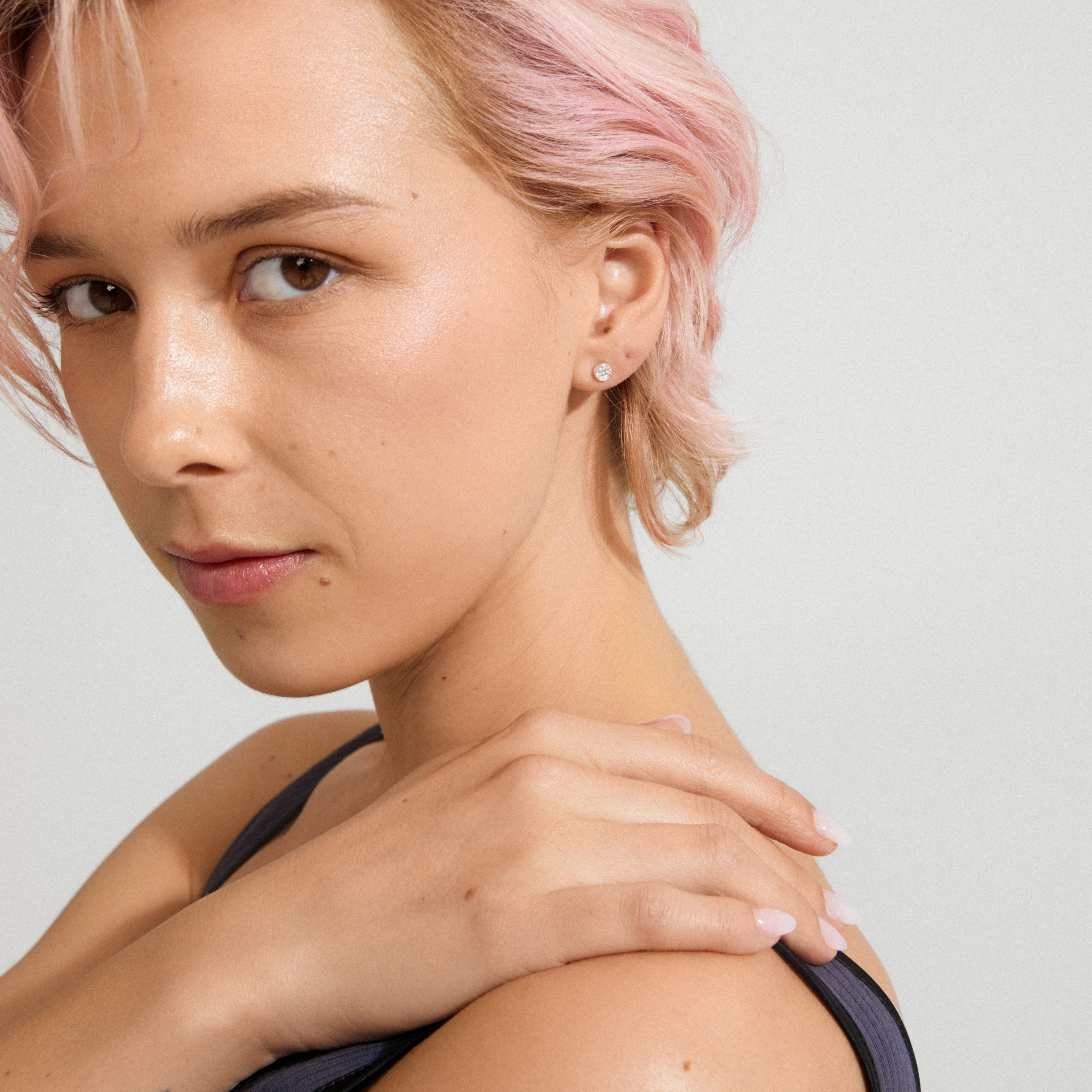 Young adult with pastel-pink hair wearing EMMA crystal earrings, silver-plated, featuring clear crystals. Hand rests on shoulder, showcasing minimalist, sparkling design amidst a stylish look.